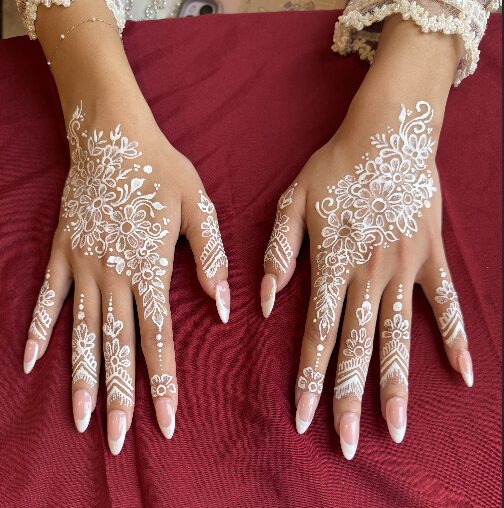 Hands with intricate white henna designs resting on red fabric, long nails, and lace cuffs visible.