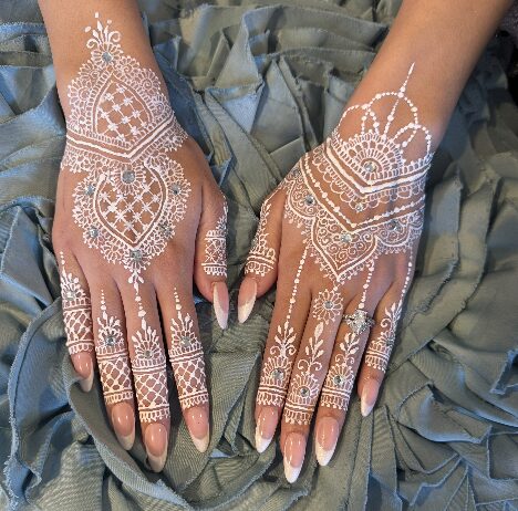Two hands with intricate white henna designs resting on gray fabric, detailed patterns cover fingers and backs of hands.