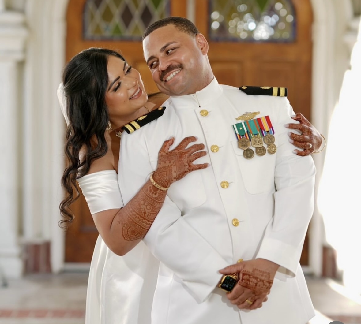 Woman hugging man in a white military uniform with medals, smiling warmly indoors.