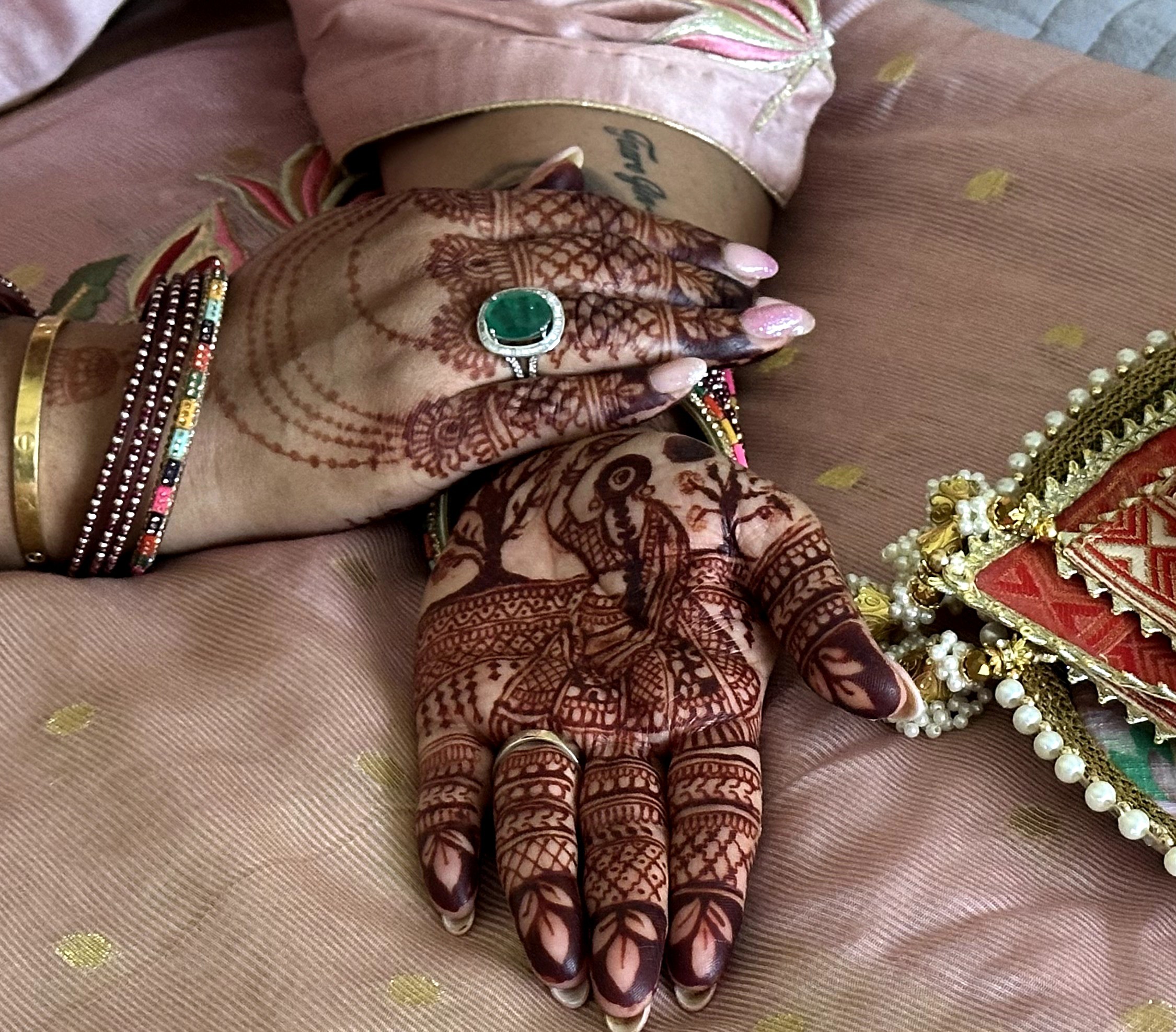 Hands with henna designs, jewelry, and a ring, resting on pink fabric with a decorative item nearby.