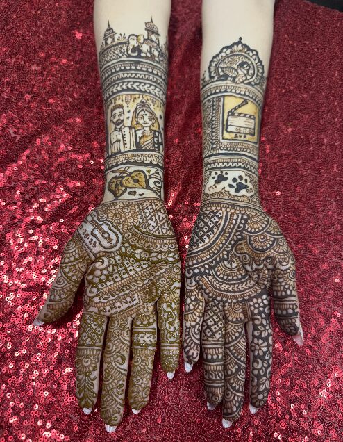 Two hands with intricate henna designs resting on a red, glittery surface, displaying detailed patterns and motifs.