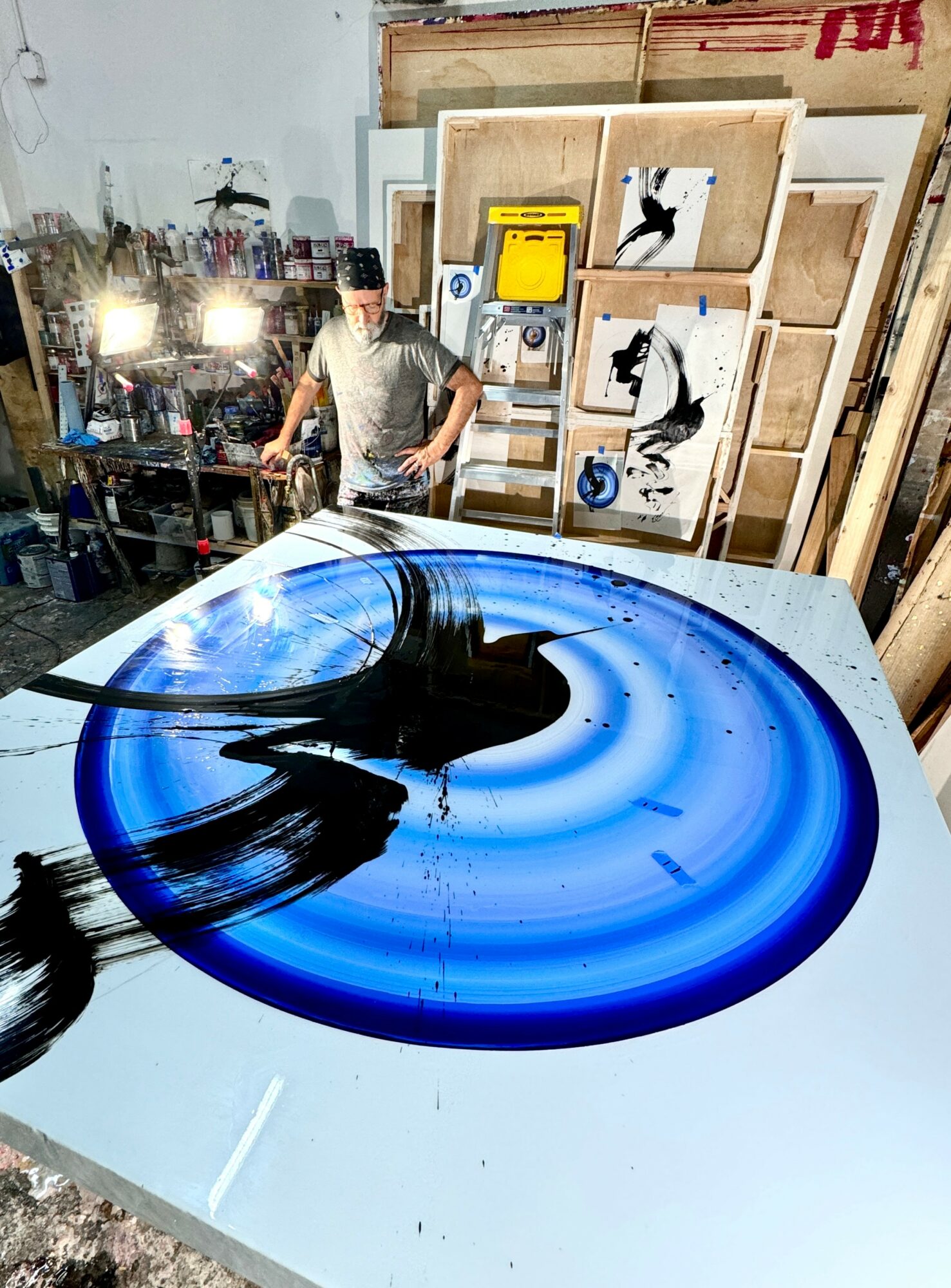 Workshop with a person standing near artwork and tools, a large circular blue and black resin art piece on the table.