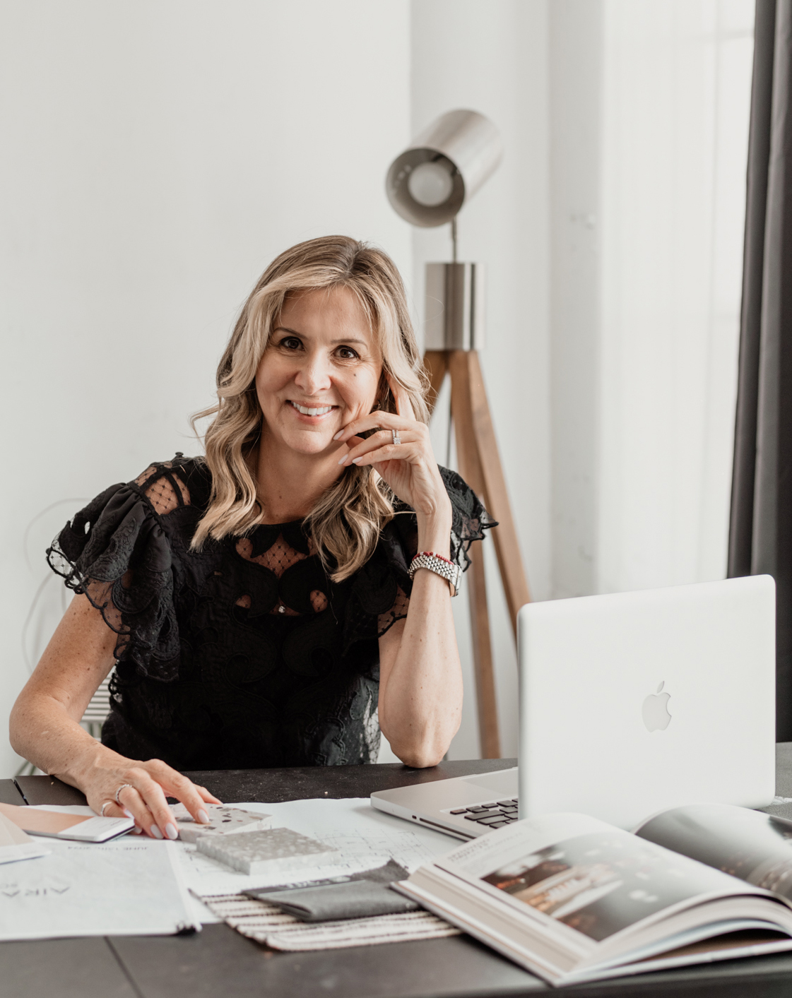 Woman sitting at desk with open laptop, papers, and magazines, smiling in a bright room with a floor lamp and window.