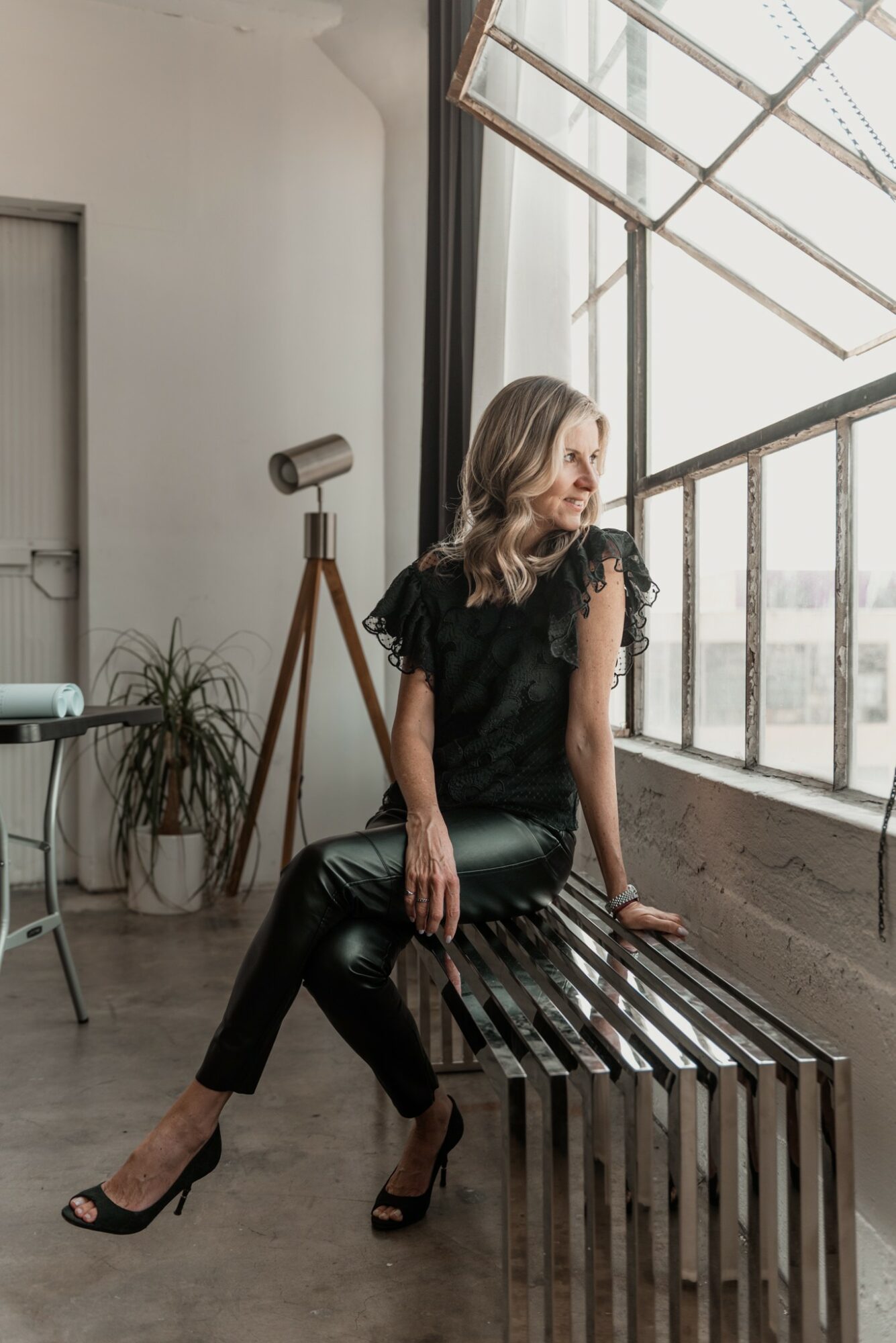Woman sitting on a wooden bench near large window with sunlight, wearing black leather pants and lace top, looking outside.