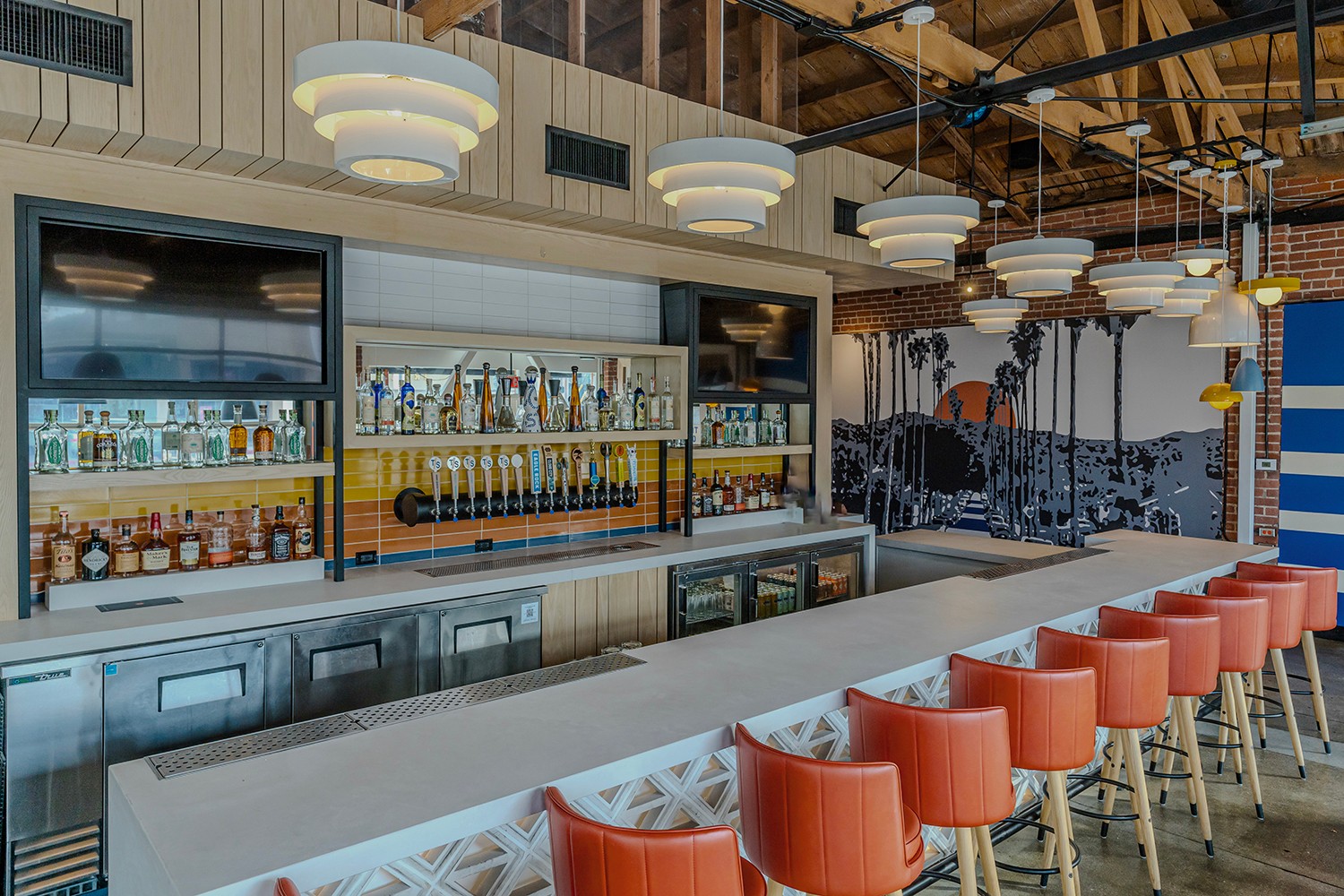 Bar area with shelves of bottles, a long counter, and orange chairs along a white table, with hanging lights and a mural.
