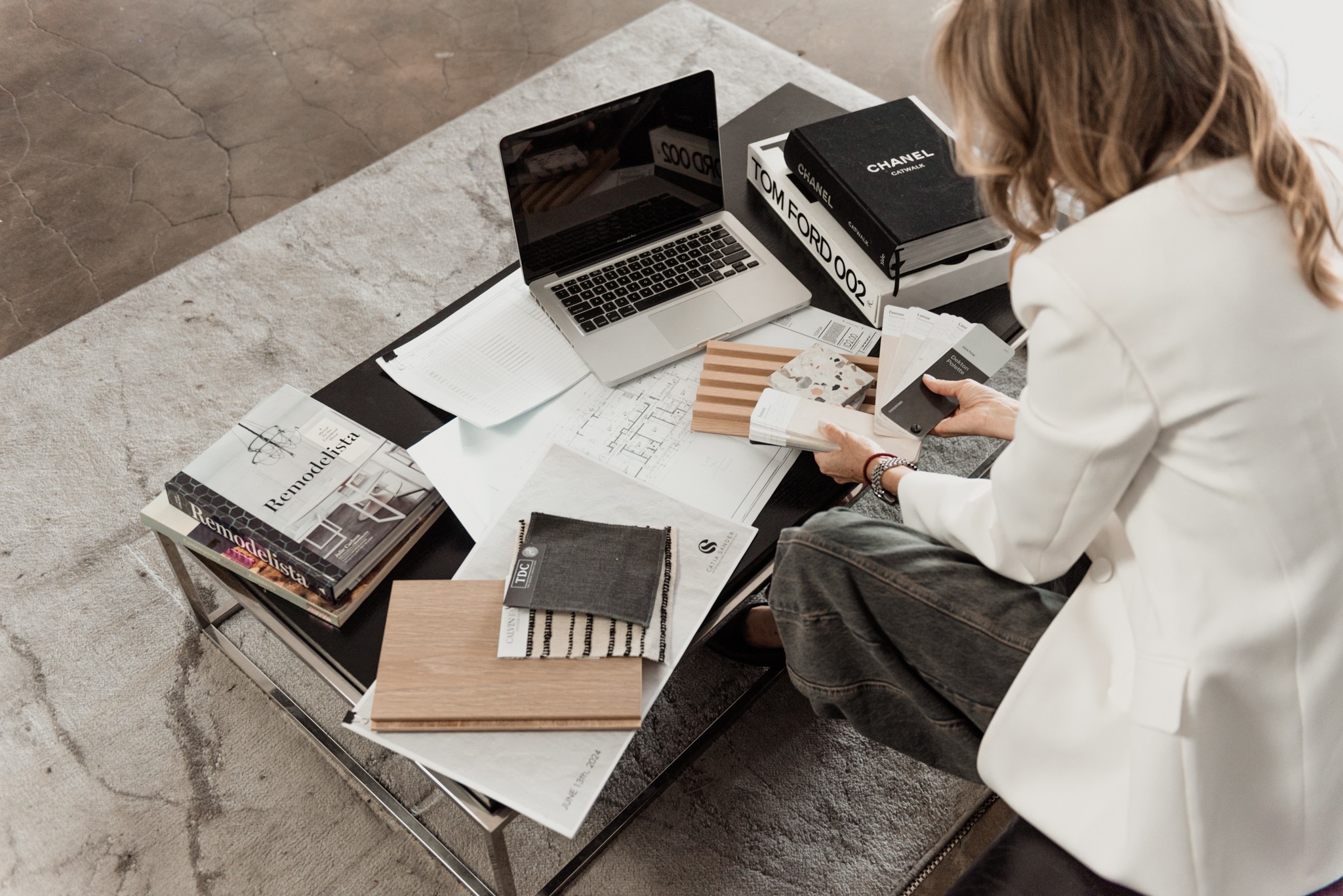 Person sitting at a table with a laptop, books, papers, and fabric samples, working on a project.