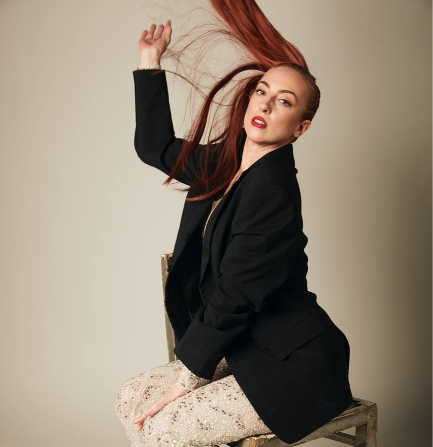 Woman with long red hair wearing a black blazer, sitting on a chair, looking at the camera.
