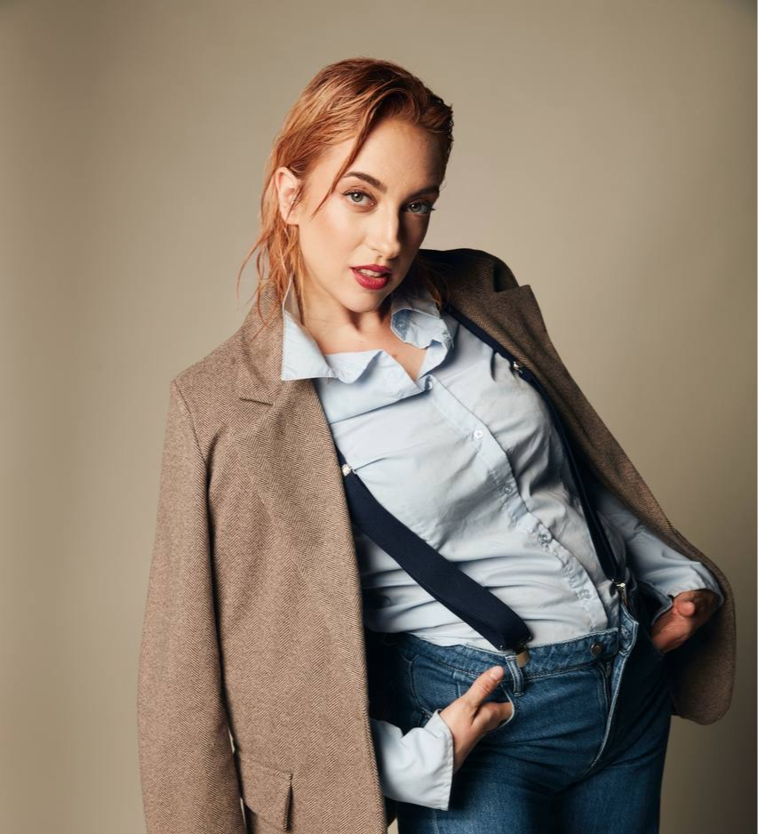 Young woman with red hair wearing a light blue shirt, dark suspenders, and a beige blazer, posing against a plain background.