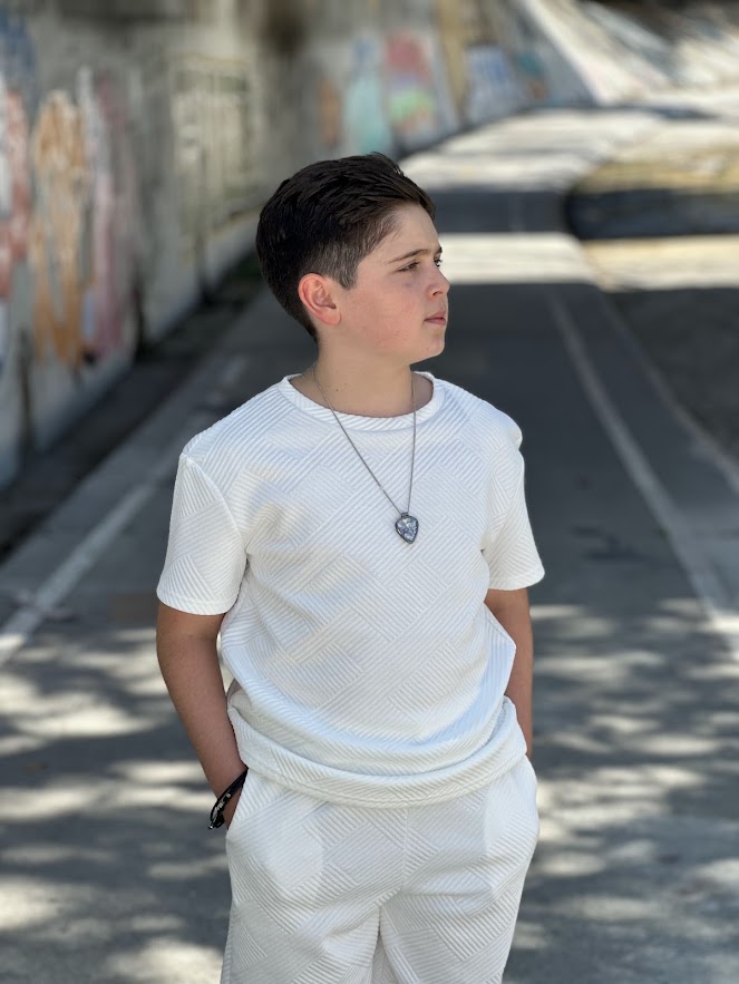 Young boy with dark hair in a white outfit standing outdoors near a graffiti wall.