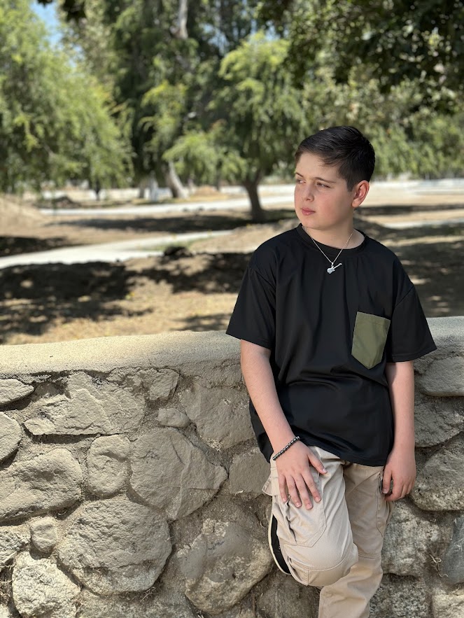Young boy leaning against a stone wall outdoors, wearing a black t-shirt and beige pants, with trees in background.
