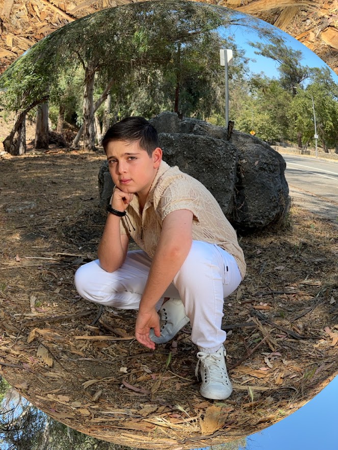 Young man crouching outdoors near a large rock, trees, and a reflective curved surface behind him.