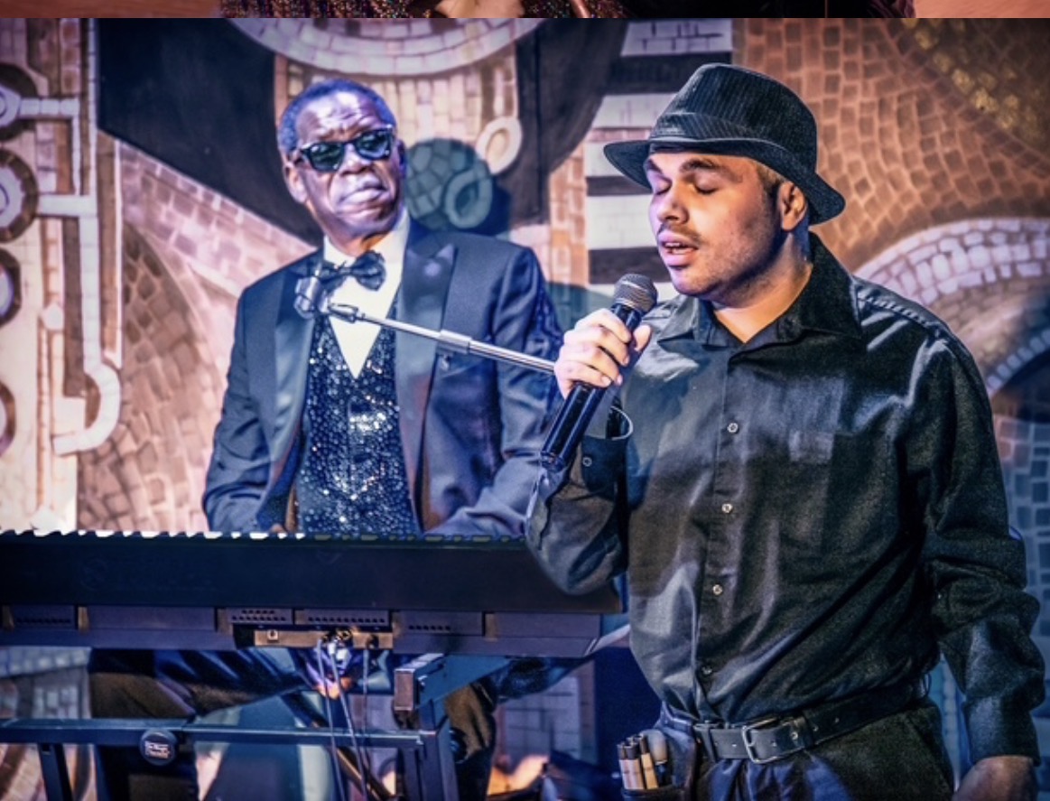 Two men performing on stage, one singing into a microphone, the other playing keyboard, with colorful background decorations.