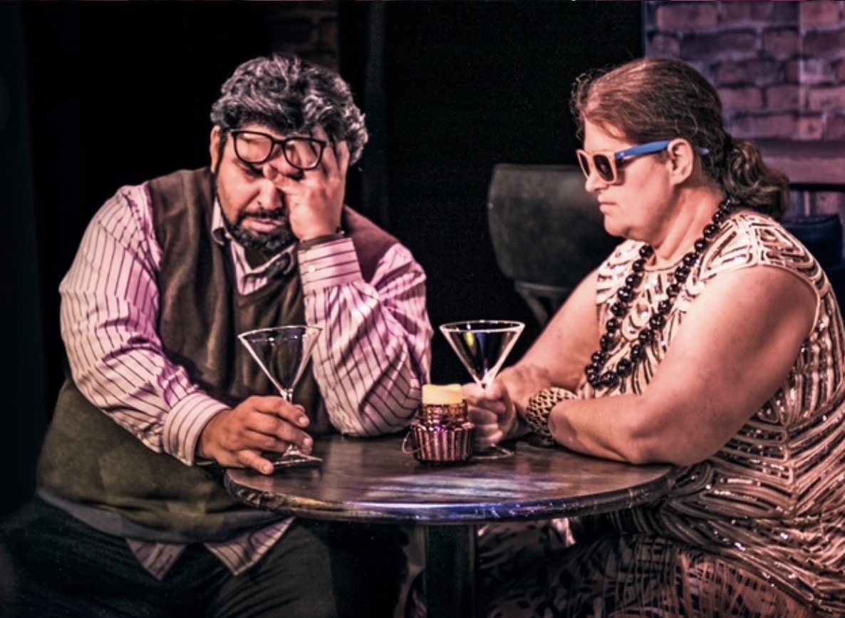Two people sit at a table with drinks, one with glasses and the other with sunglasses, in a dimly lit setting.