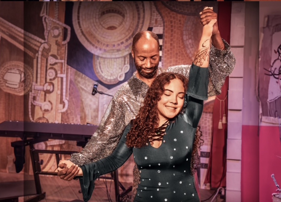 A smiling man and woman dance together on stage, holding hands, with colorful abstract background and stage equipment.