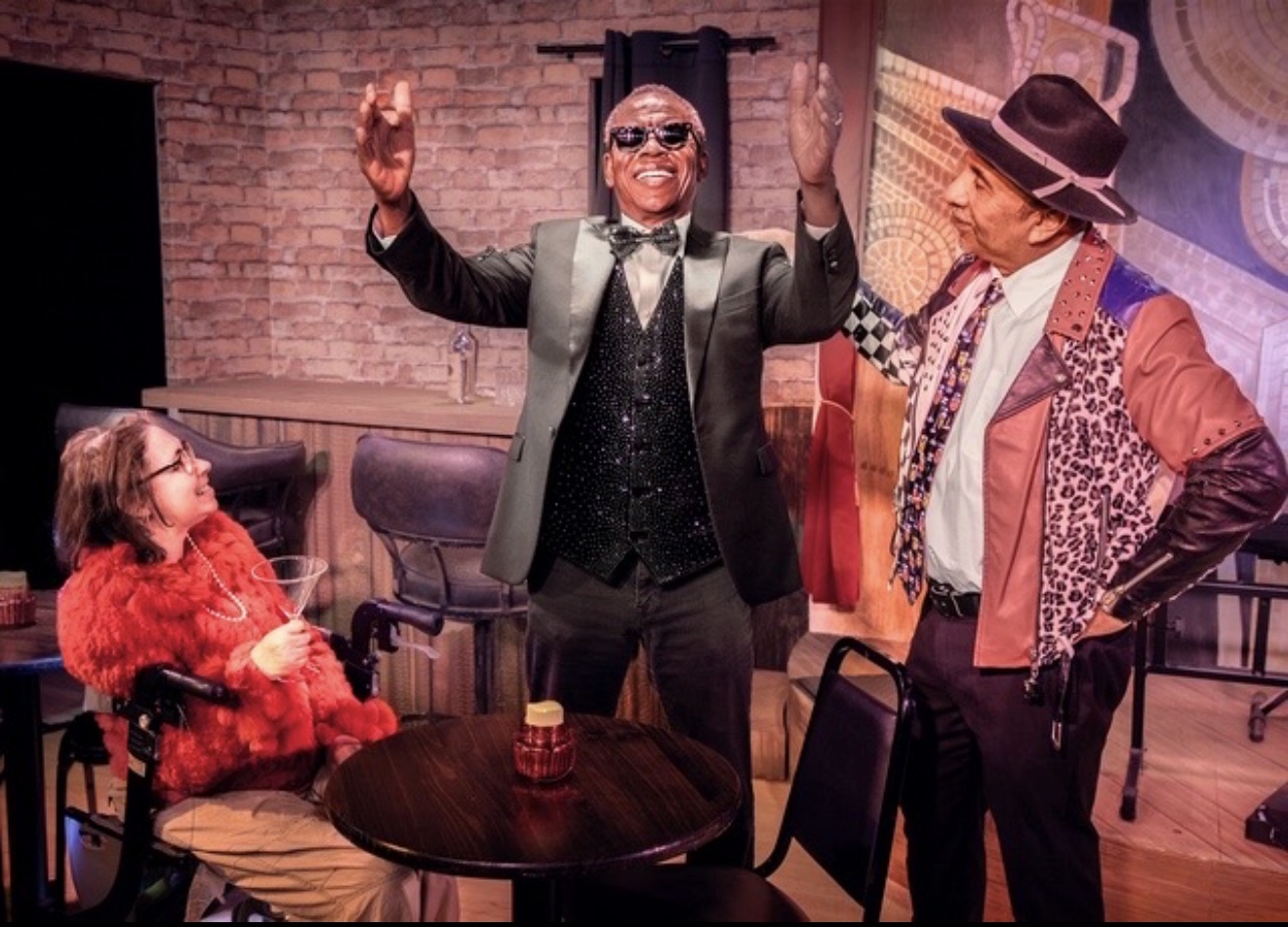 Three people on stage, one sitting and two standing, with a brick wall background and a table with a candle.