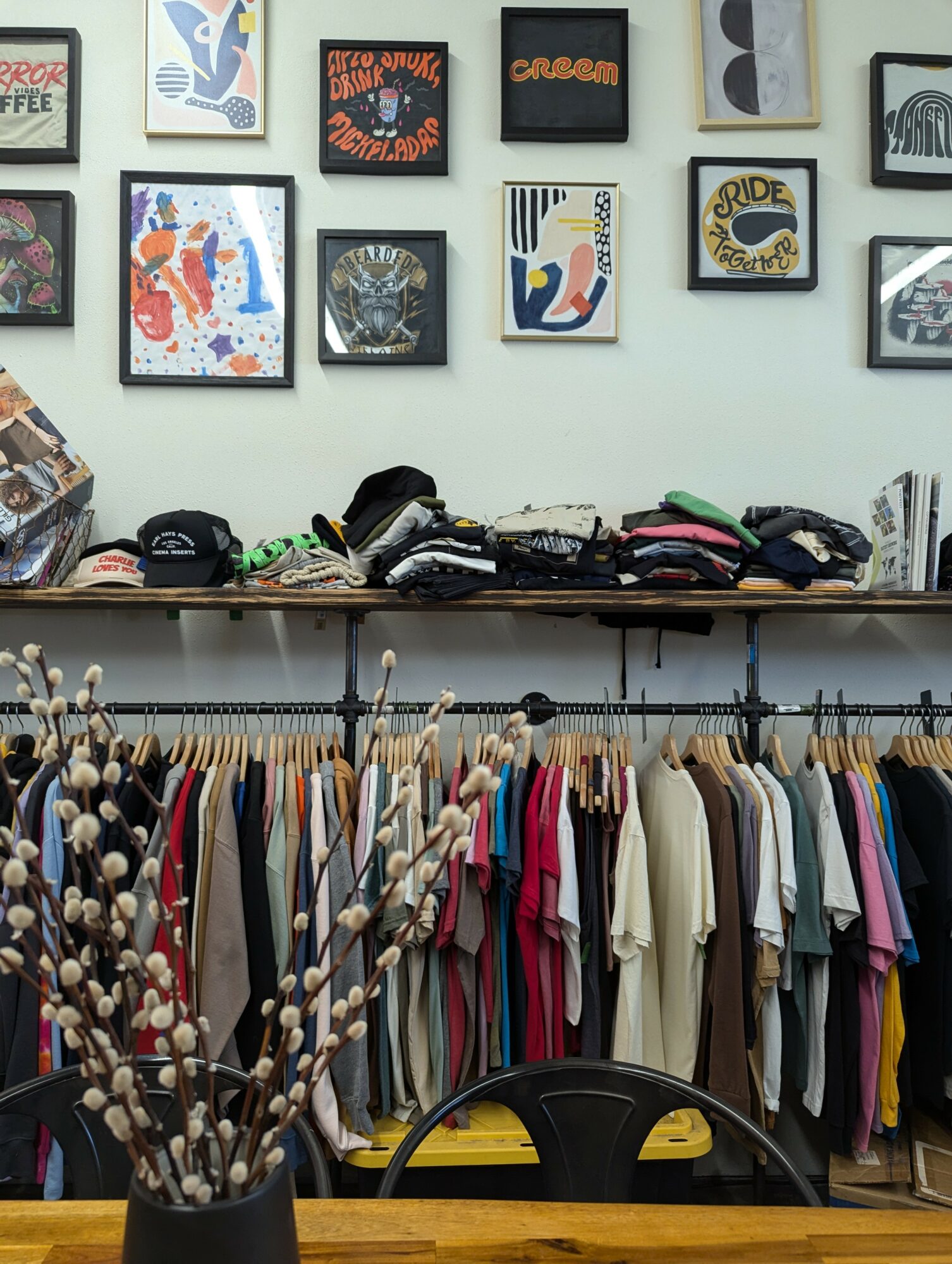 Clothing rack with colorful shirts and jackets, wall with framed artwork, and a table with folded clothes and hats.