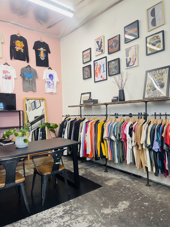 Clothing store interior with framed artwork on wall, clothing rack with colorful shirts, table with chairs, and pink wall with T-shirts.