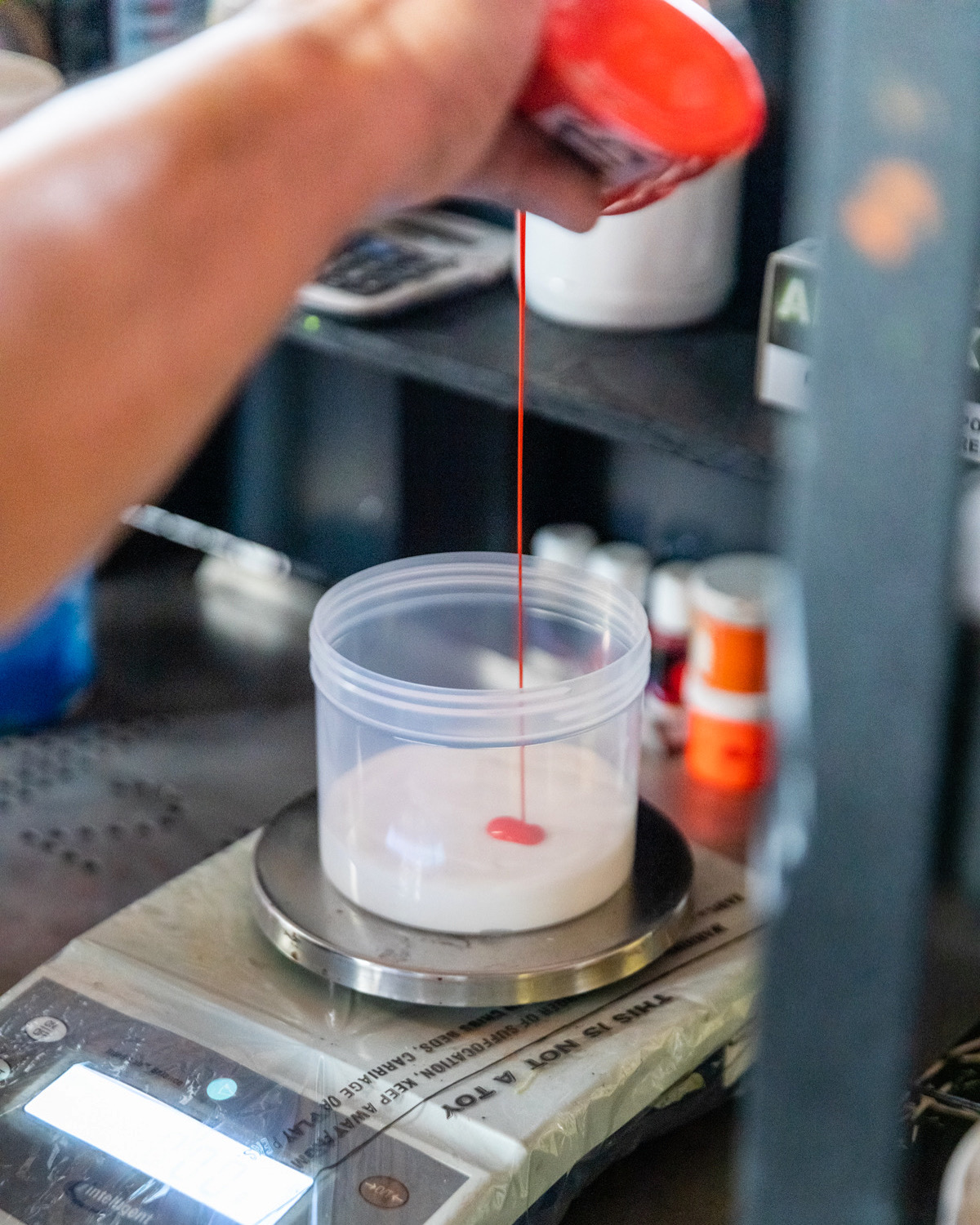 Person pouring red liquid into a clear container on a digital scale, with shelves and bottles in background.