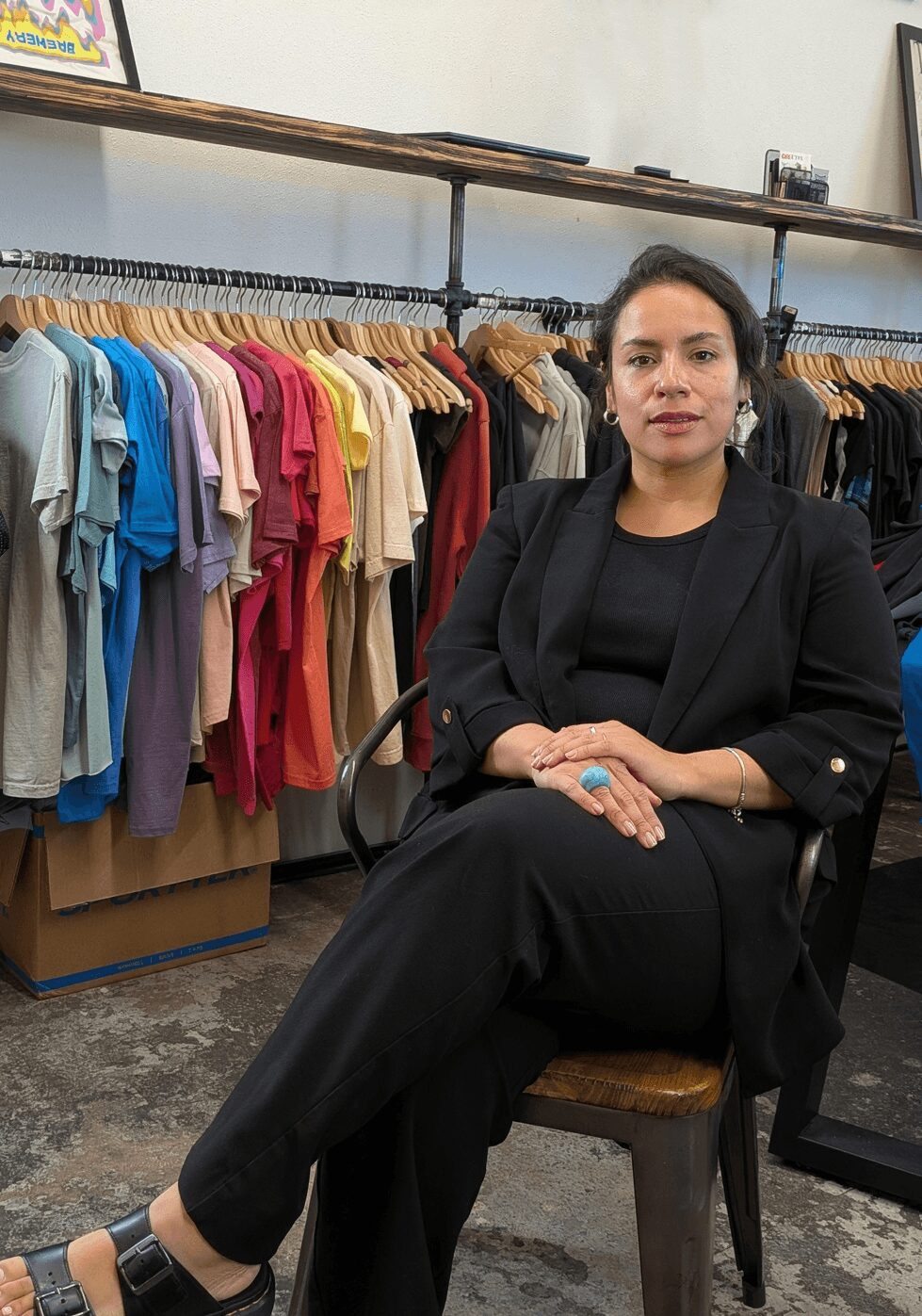 Woman sitting on a chair in a clothing store with racks of colorful shirts behind her.