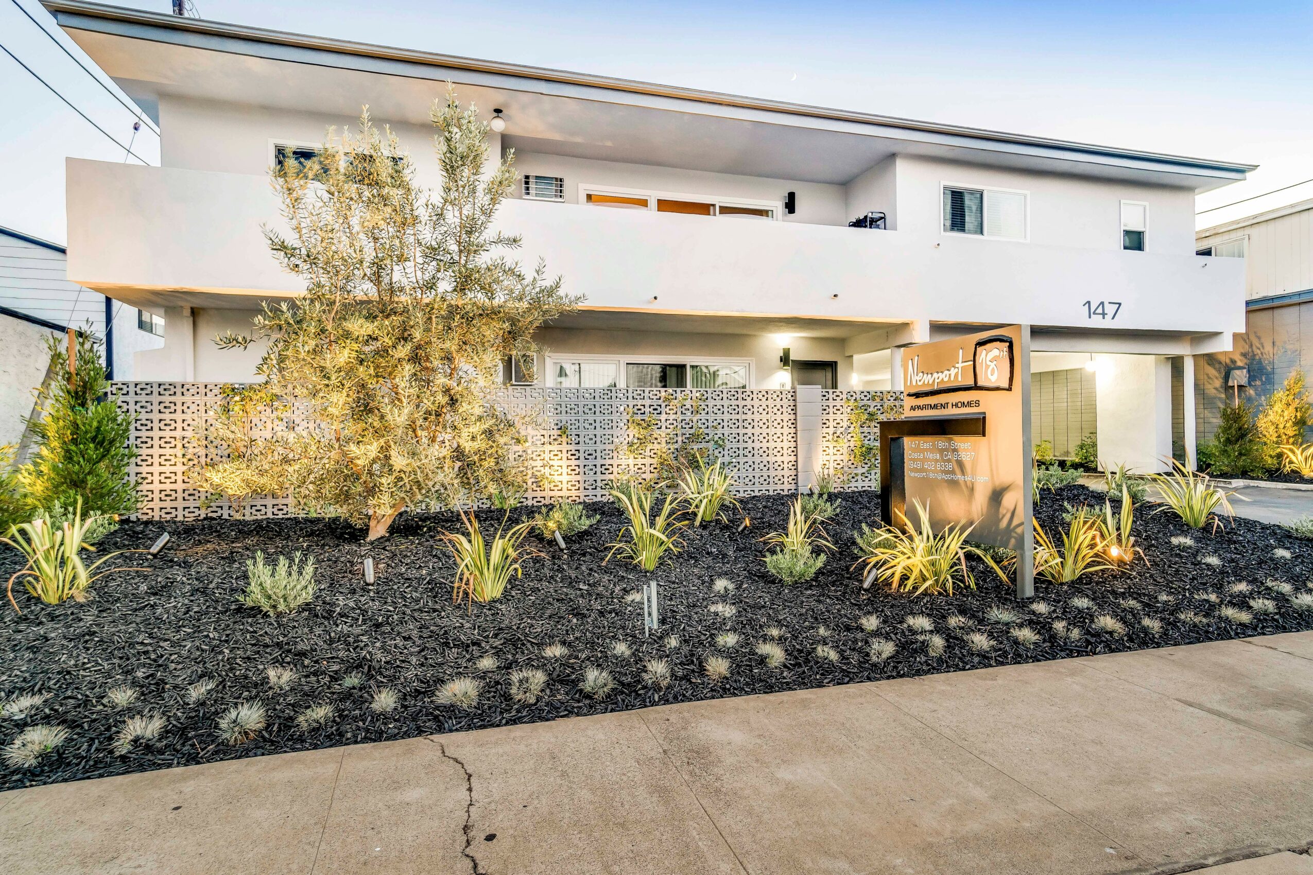 Modern white building with balcony, surrounded by landscaped garden with plants and mulch, mailbox near sidewalk.