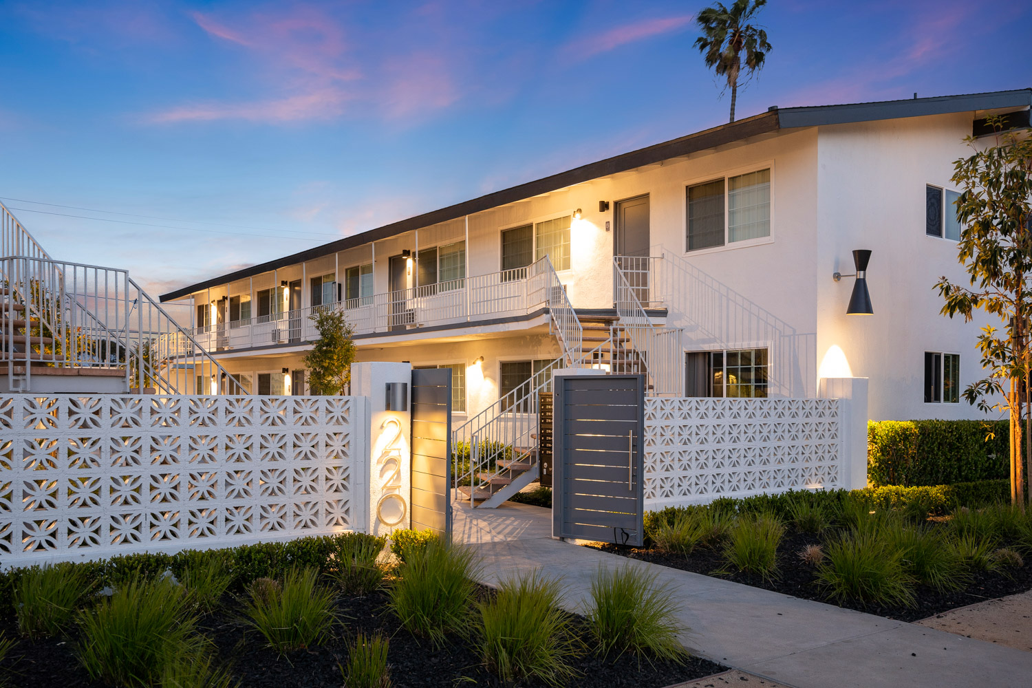 Two-story white apartment building with balconies, surrounded by a decorative wall and landscaped garden, under a colorful evening sky.