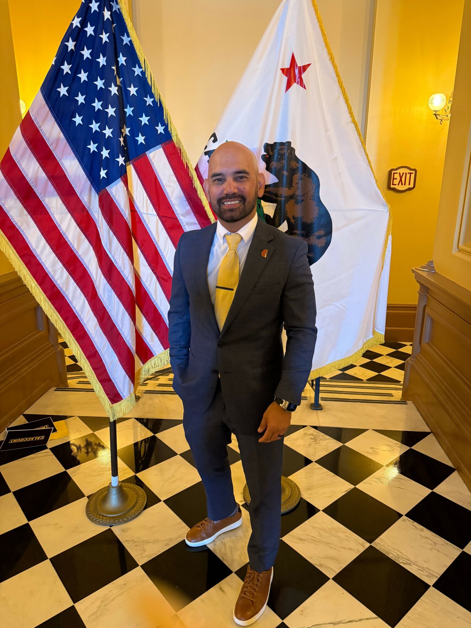 Man in suit standing in front of American and California flags indoors, smiling, with black and white checkered floor.