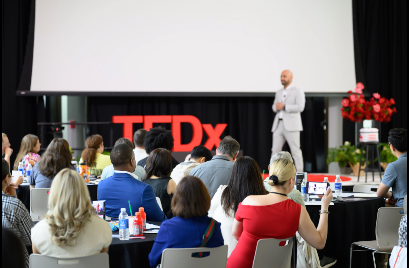 Speaker on stage with TEDx sign, audience seated, some taking photos, flowers on stage, large blank screen behind.