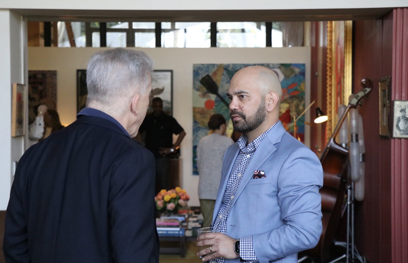 Two men engaged in conversation indoors, with artwork and people in the background.