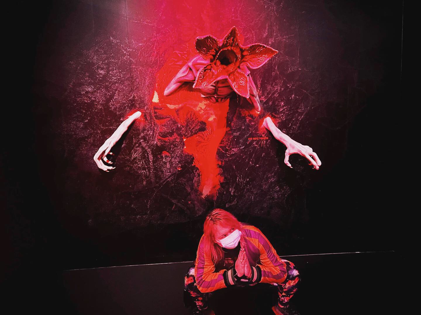 Person wearing a mask and red clothing crouching with hands clasped, in front of a dark background with a red figure above.