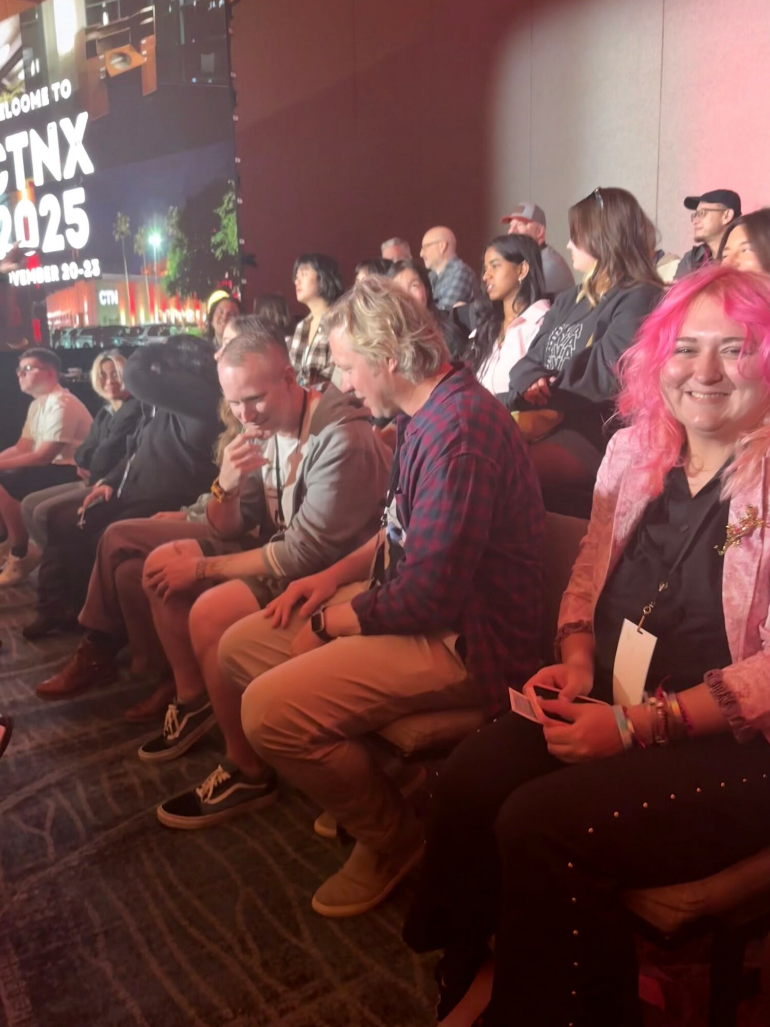 Group of people seated in a dark room, some looking at their phones, others talking or smiling.