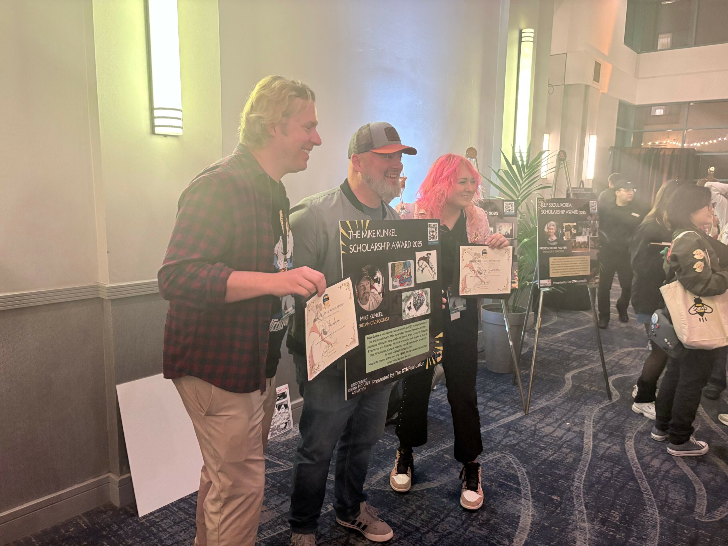 Three people holding certificates and standing indoors near display boards, with a group of people in the background.