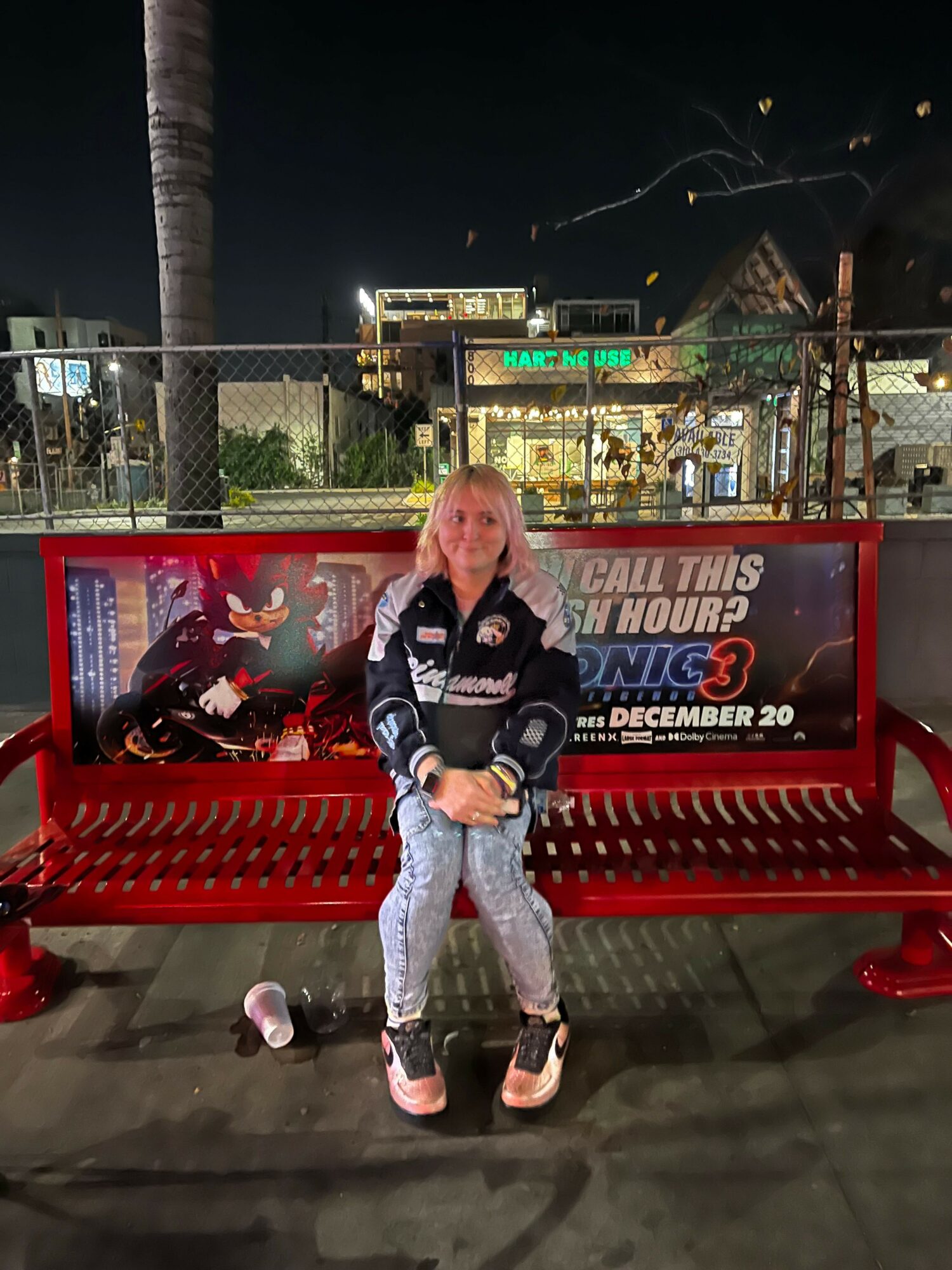 Woman sitting on a red bench at night, with a motorcycle advertisement behind her, palm trees, and buildings in the background.