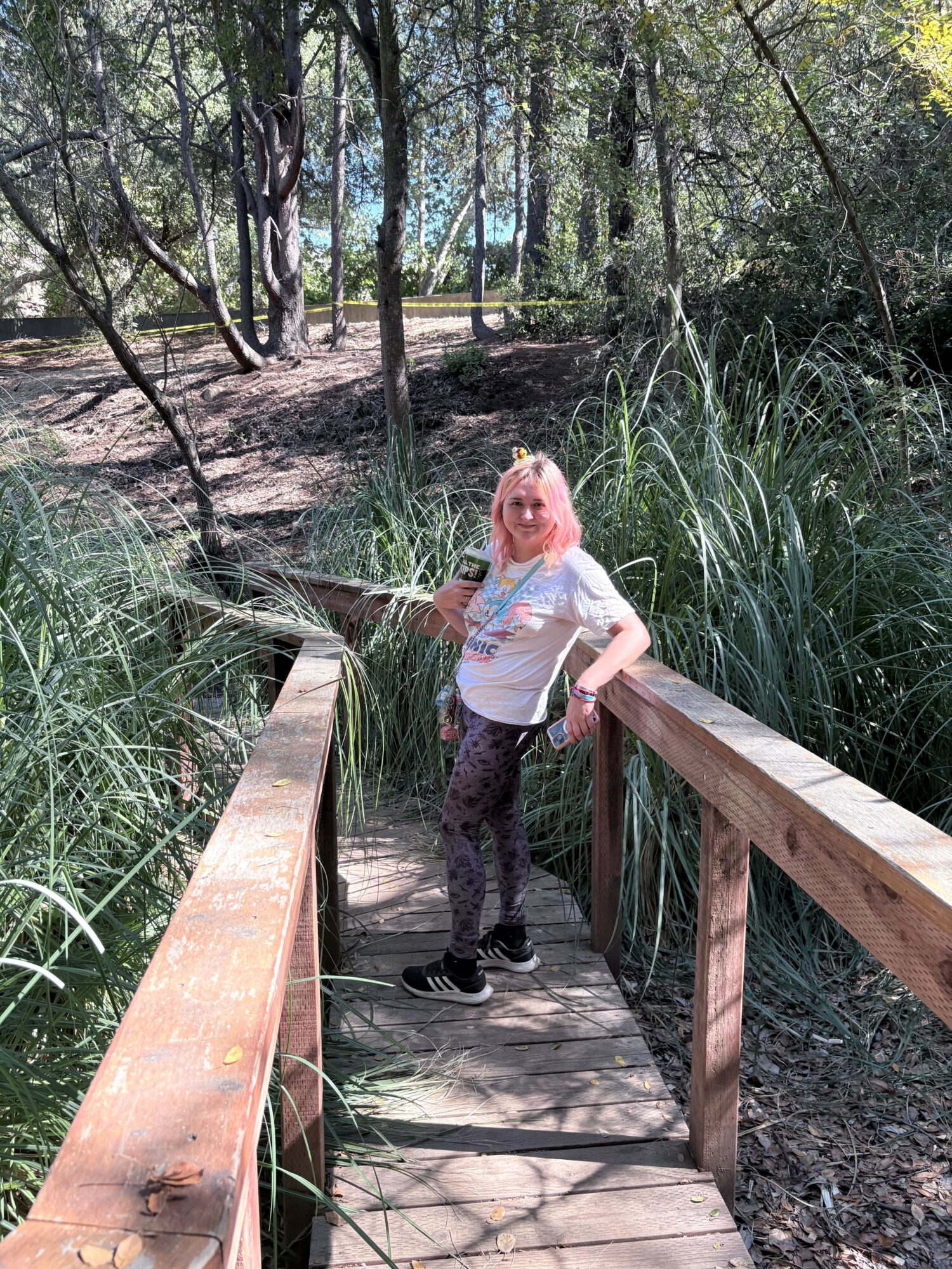Woman with blonde hair standing on a wooden bridge in a forested area, surrounded by tall grass and trees.