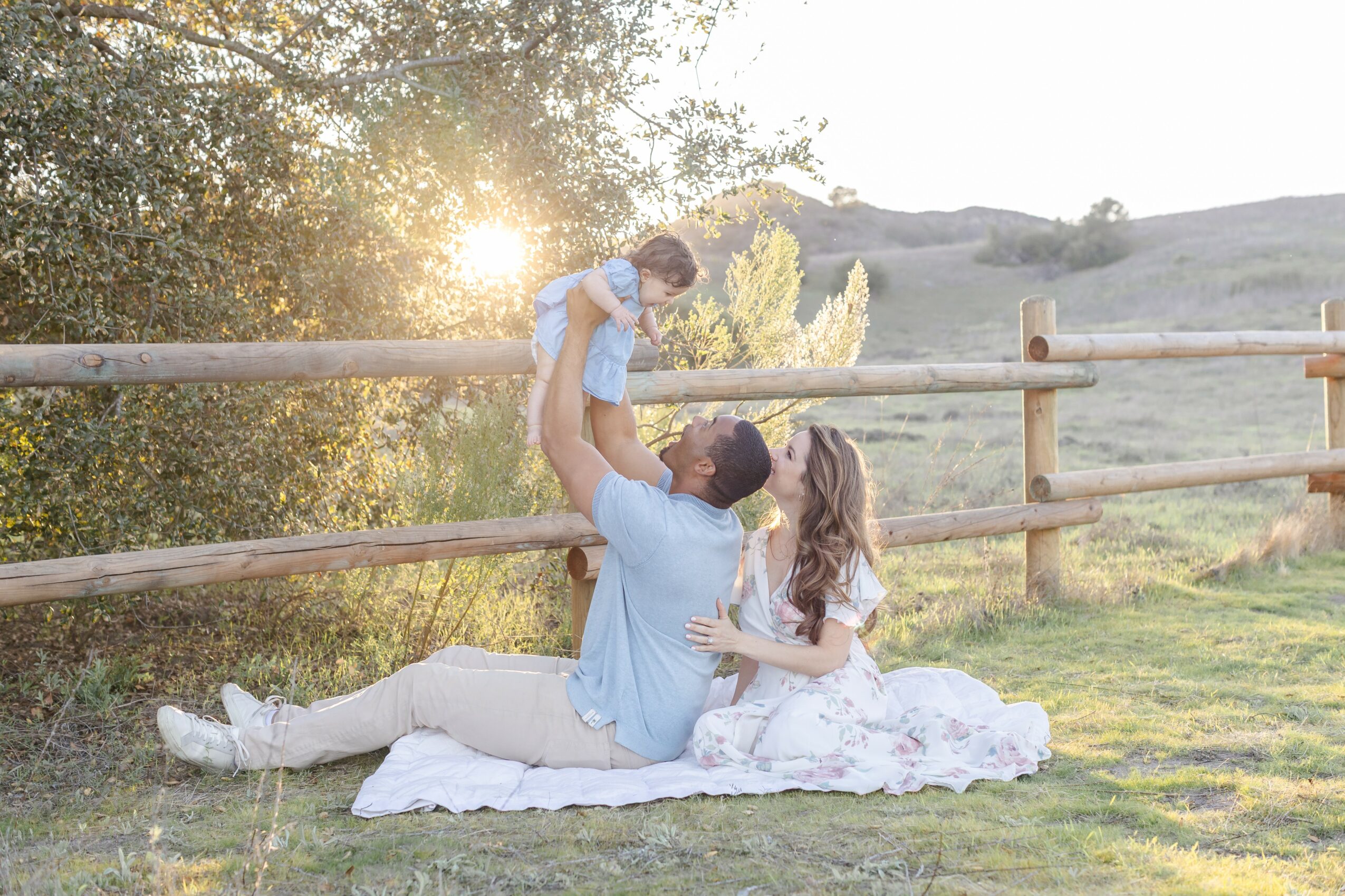 Family of three outdoors on a blanket, with a man lifting a child, a woman watching, and a wooden fence nearby.
