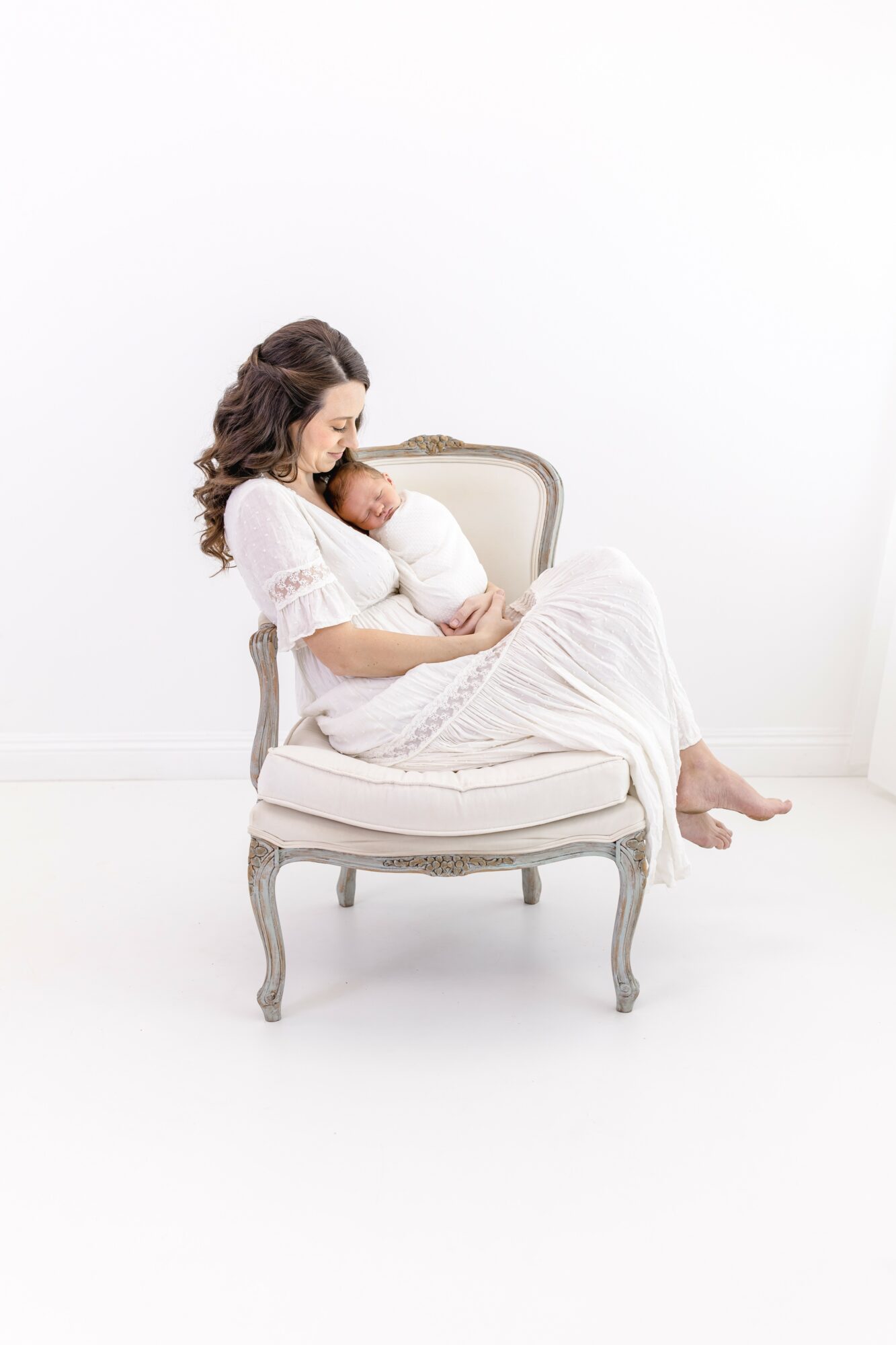Woman sitting on a vintage chair holding a baby, both wearing white dresses, against a plain white background.
