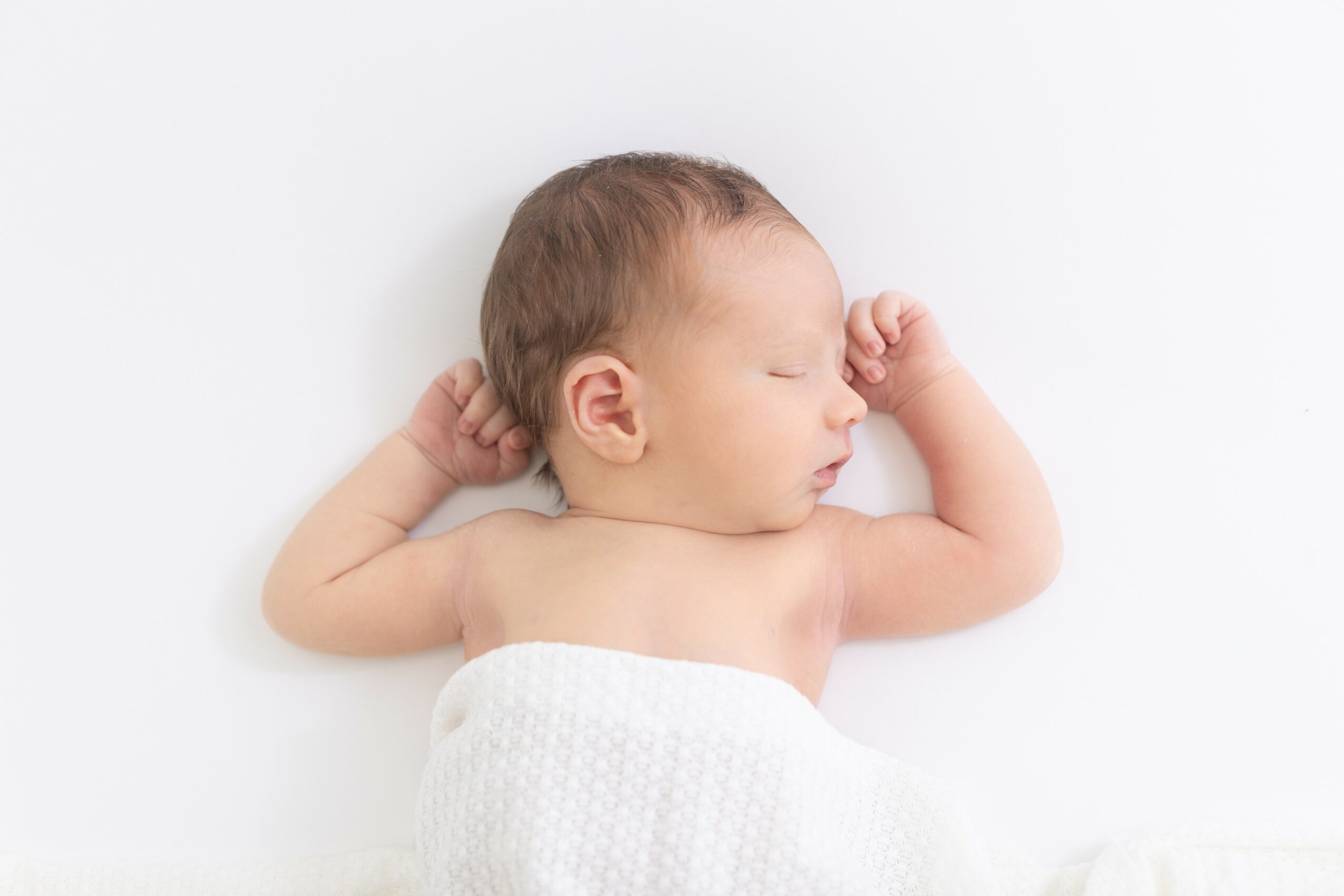 Baby sleeping on white surface, wrapped in a white towel, with arms raised near head.