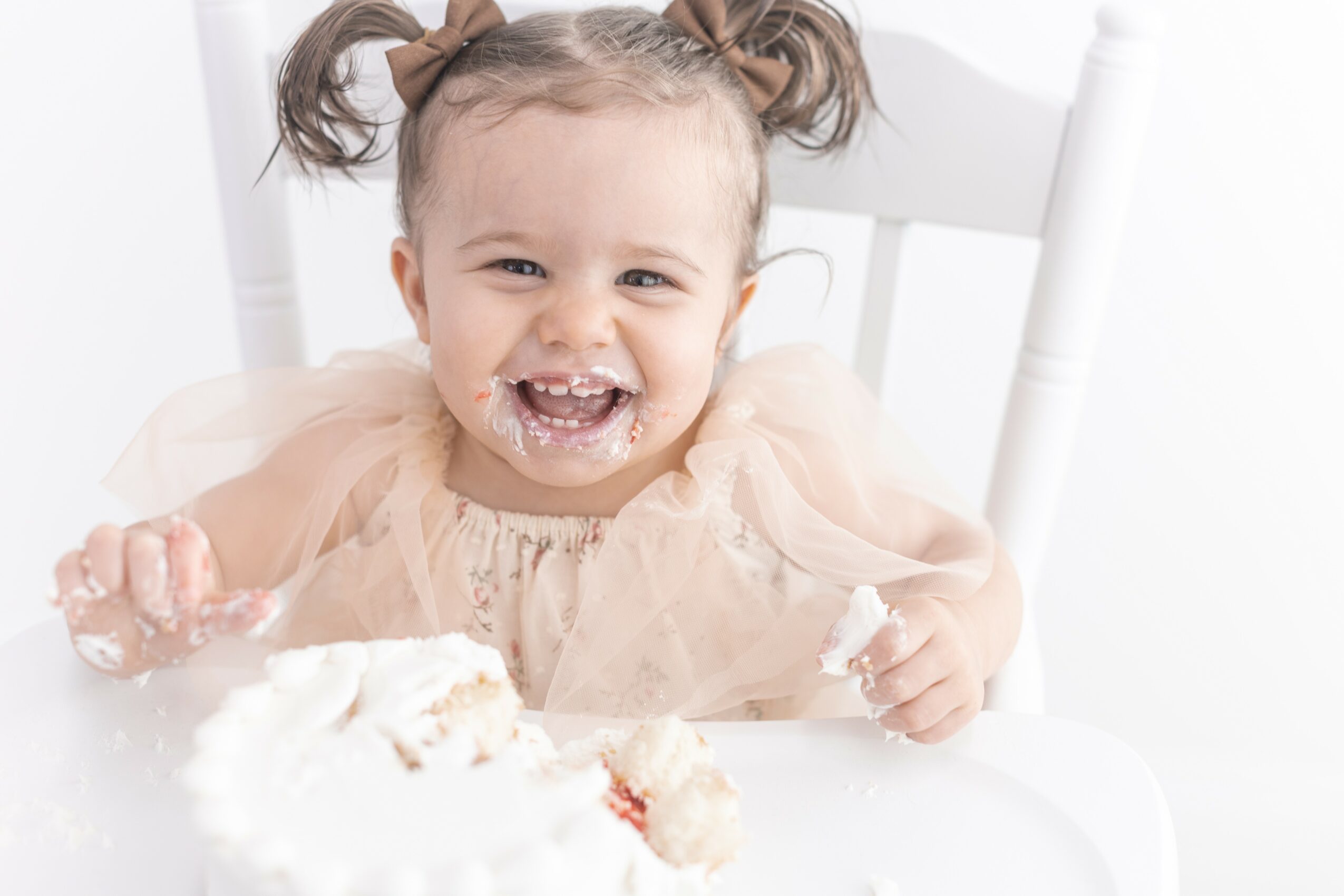 Child with puffy hair and a dress, smiling with cake on face, sitting in a white high chair.