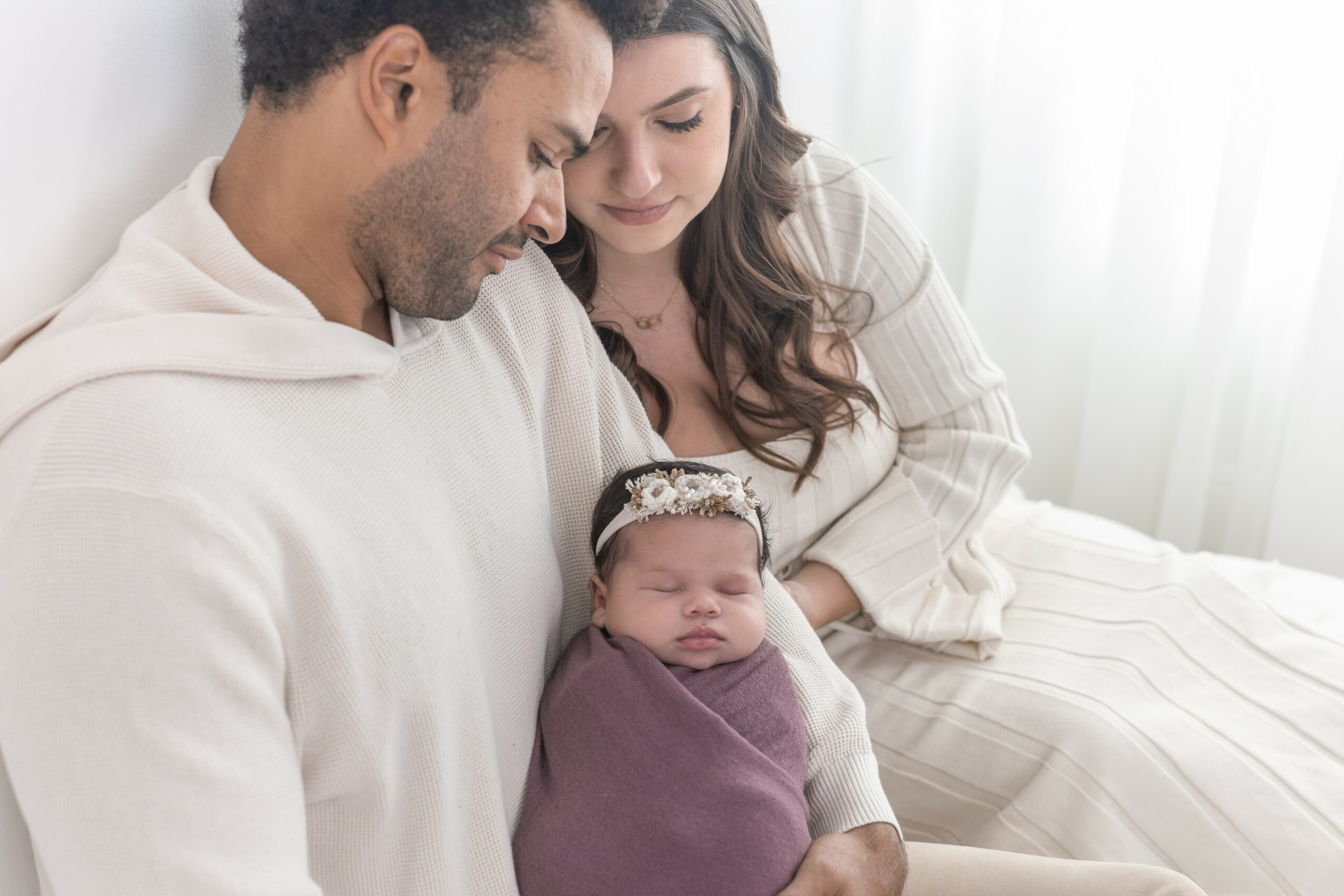 Family with a newborn baby sitting on a bed, parents looking at the baby, bright room with curtains in background.