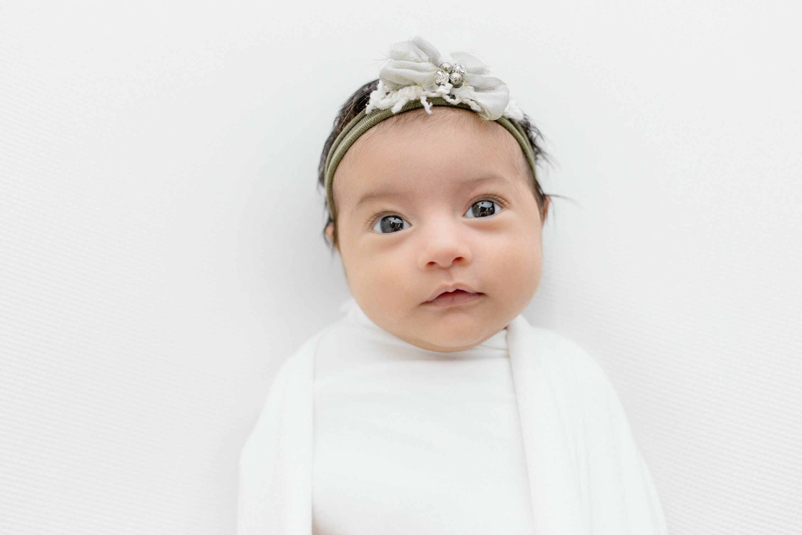Baby girl with headband and white outfit looking at camera against white background.