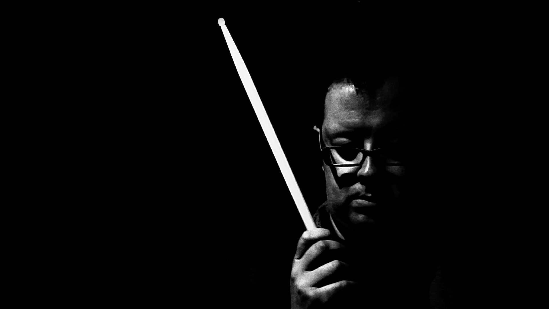 Person holding a long stick or rod, partially visible face, dark background, glasses, serious expression.