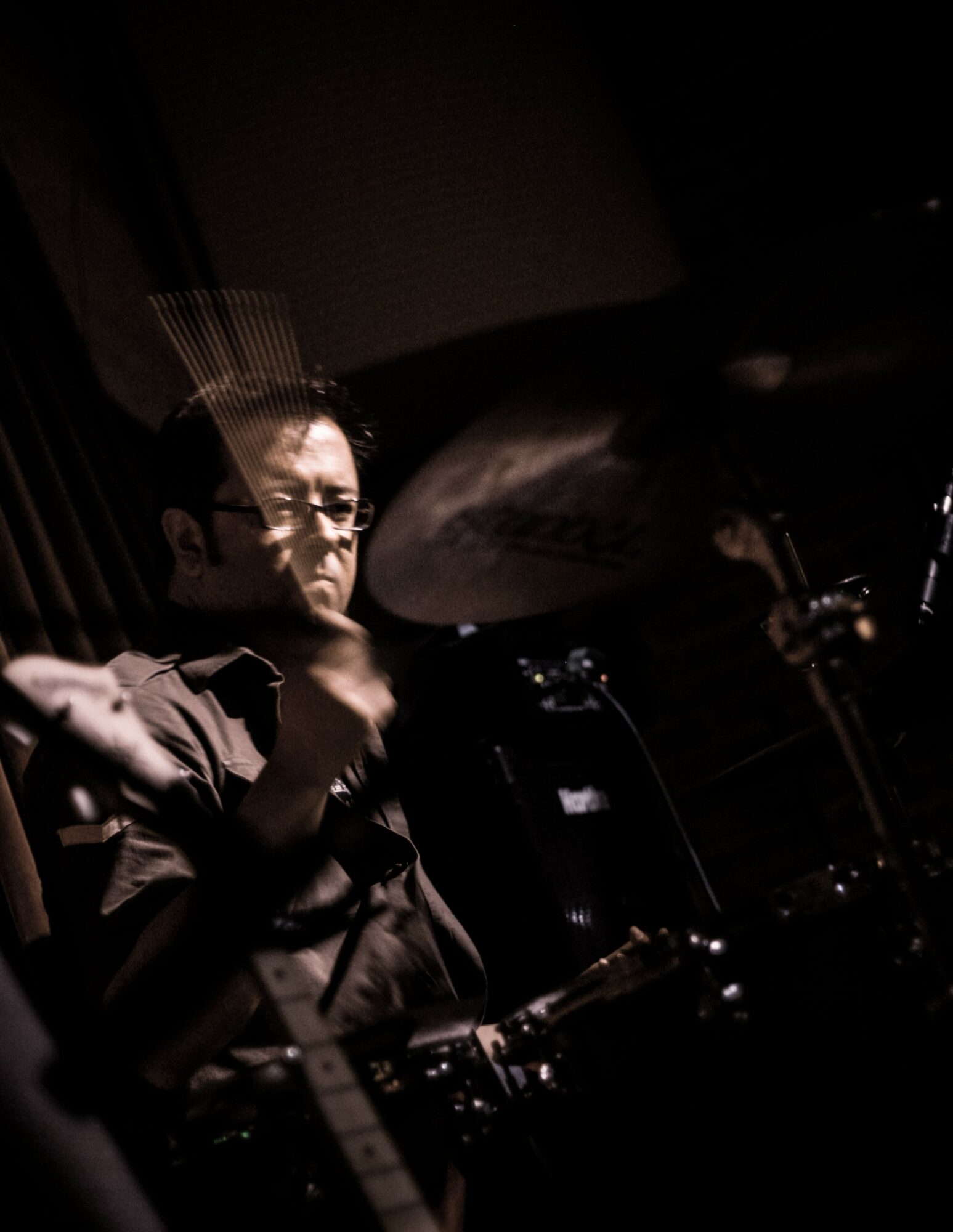 Person playing drums in a dark setting with a focused expression, glasses, and a drumstick in hand.