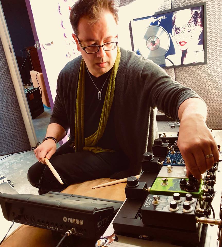 Person with glasses and short hair adjusts audio equipment in a room with computer monitors and artwork.
