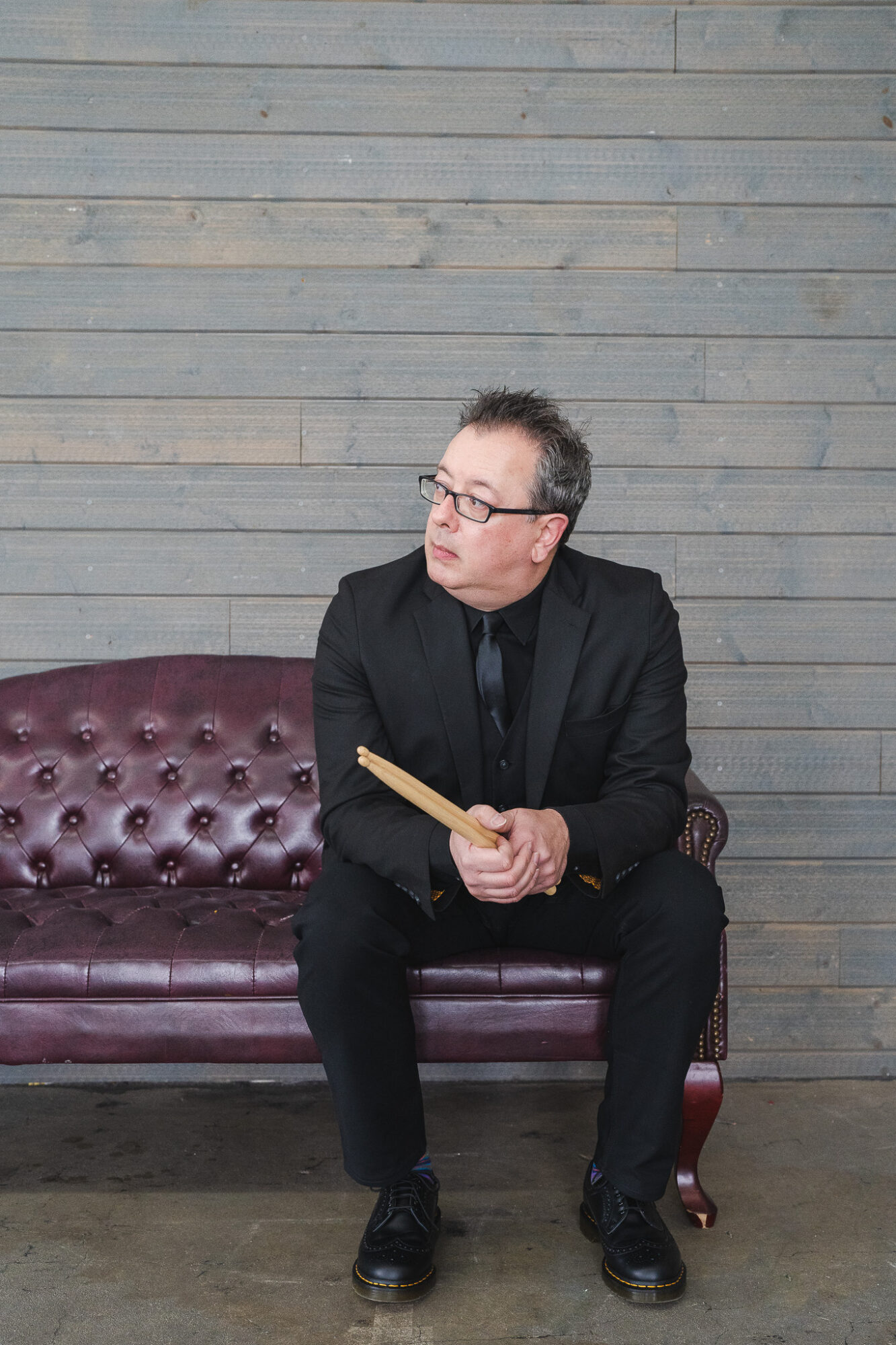 Man in black suit and glasses sitting on a purple leather couch holding a book, against a wooden wall background.