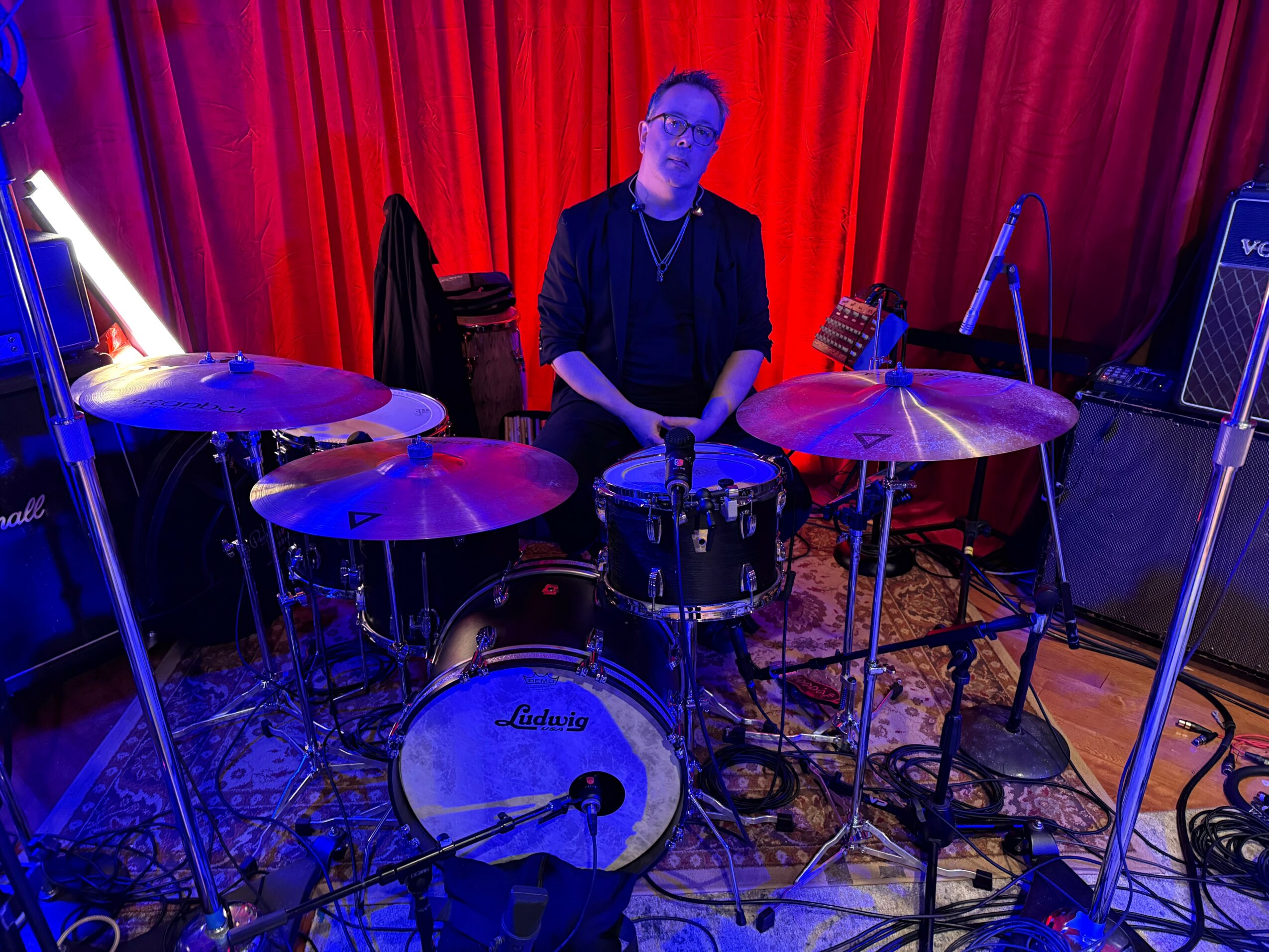 Person sitting behind a drum set with red curtains in the background, surrounded by musical equipment.