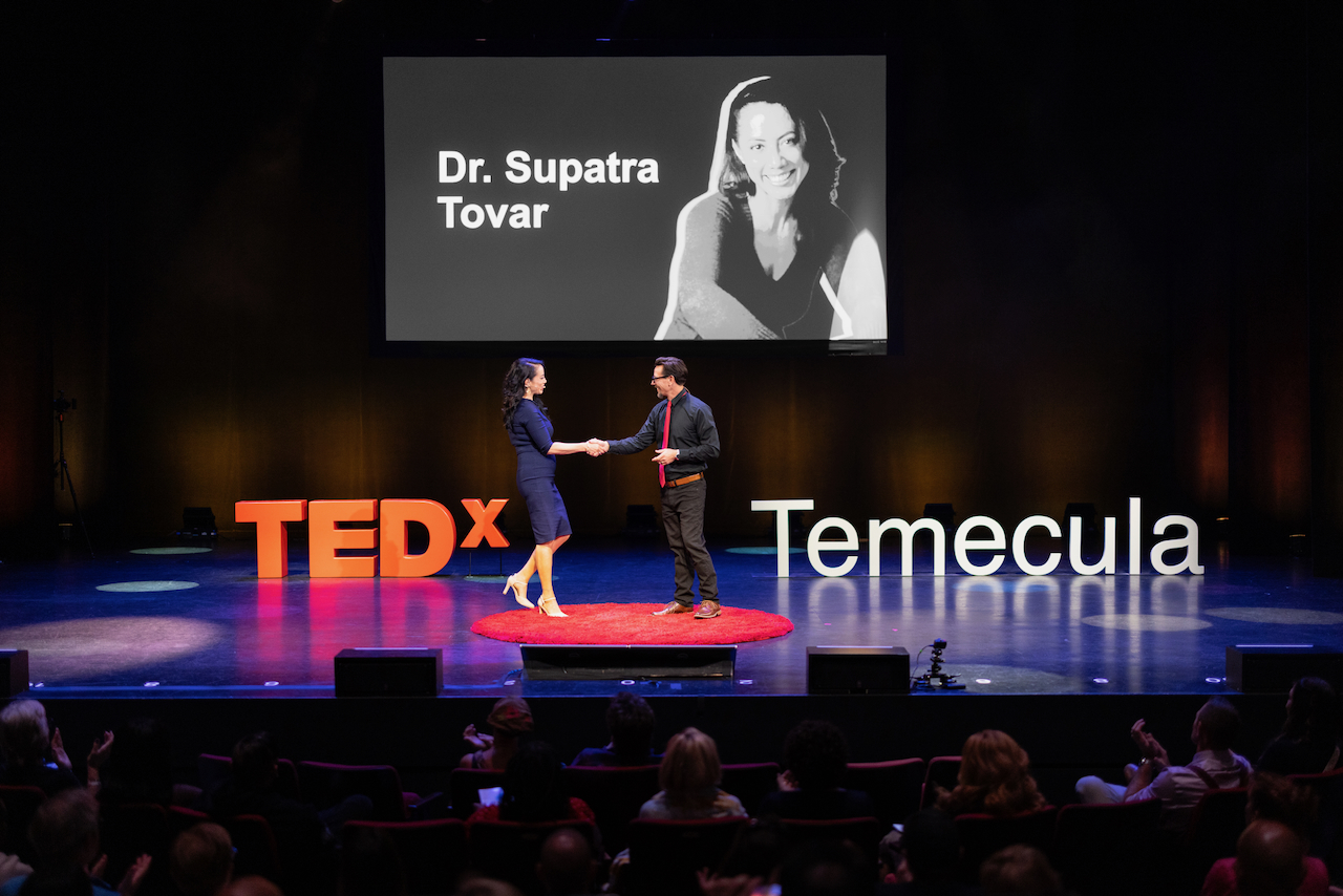 Two people on stage exchanging a handshake, with TEDx and Temecula signs, and a large screen displaying a black-and-white photo of a woman.