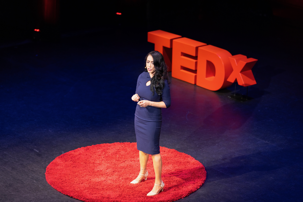 Woman standing on stage with TEDx logo in background, speaking to audience, wearing a dark dress, on a red circular carpet.