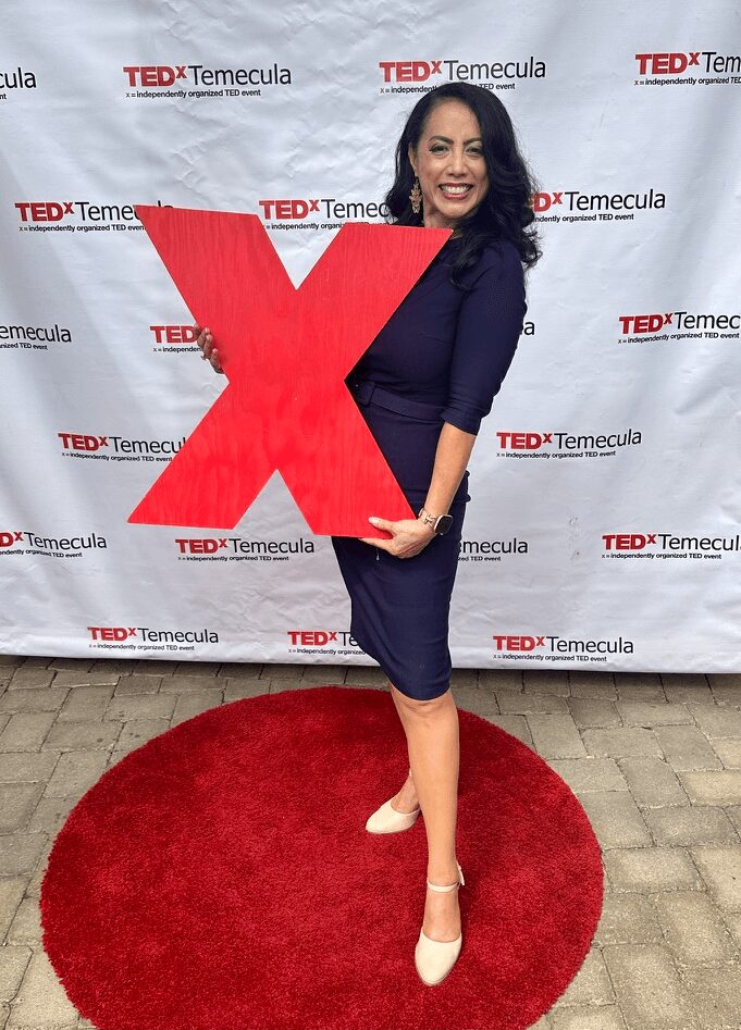 Woman in navy dress holding a large red TEDx logo sign, standing on a red carpet in front of a TEDx backdrop.
