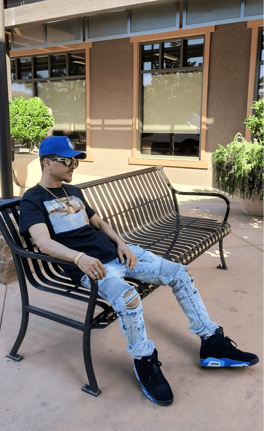 Young person wearing a blue cap, sunglasses, black t-shirt, ripped jeans, and black sneakers sitting on a park bench.