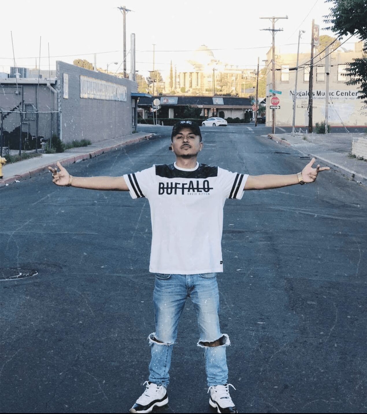 Young person standing in the middle of an empty street with arms outstretched, wearing a white t-shirt and ripped jeans.