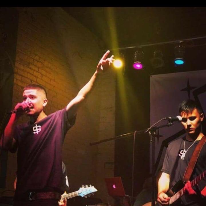 Two male performers on stage, one singing into a microphone with his arm raised, the other playing guitar, with colorful stage lights.